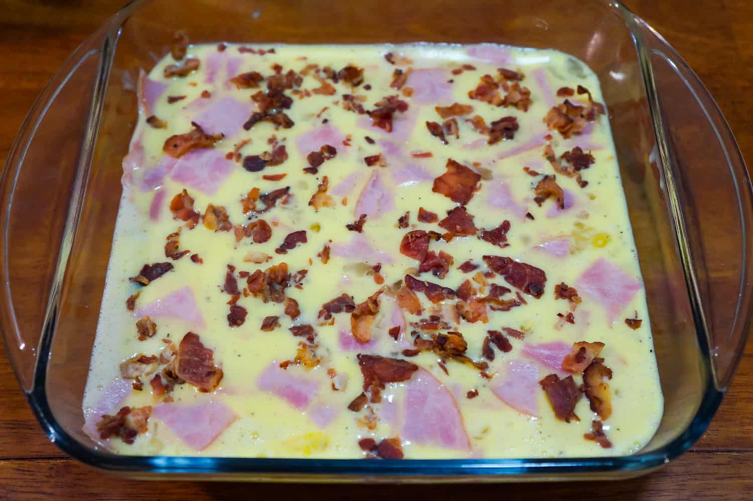 chopped ham and bacon bits on top of egg mixture in a baking dish