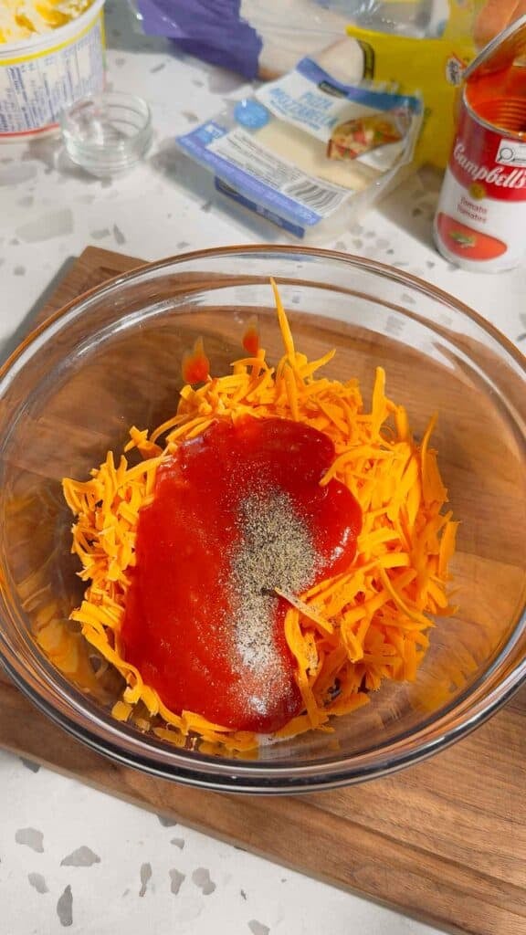 salt, pepper and condensed tomato soup on top of shredded cheddar cheese in a glass bowl