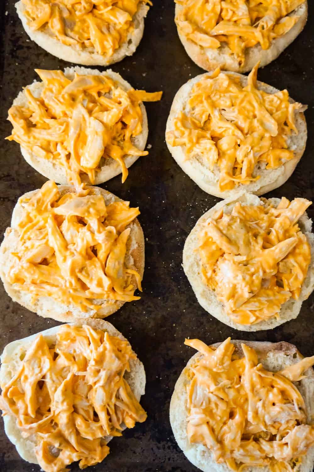 shredded chicken coated in Buffalo sauce on top of English muffins on a baking sheet