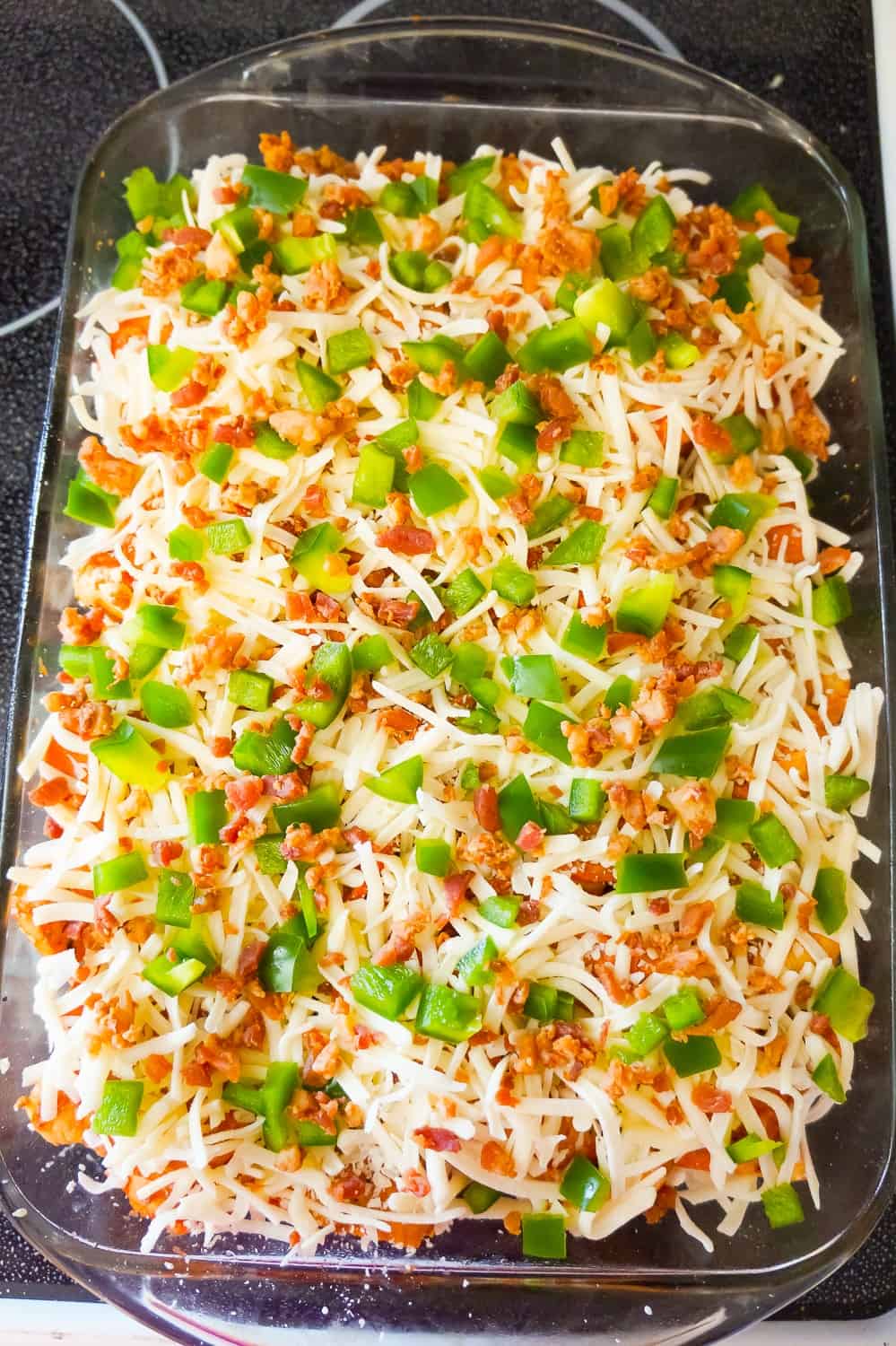 pizza bagel casserole topped with shredded mozzarella, diced green peppers and bacon crumble before baking