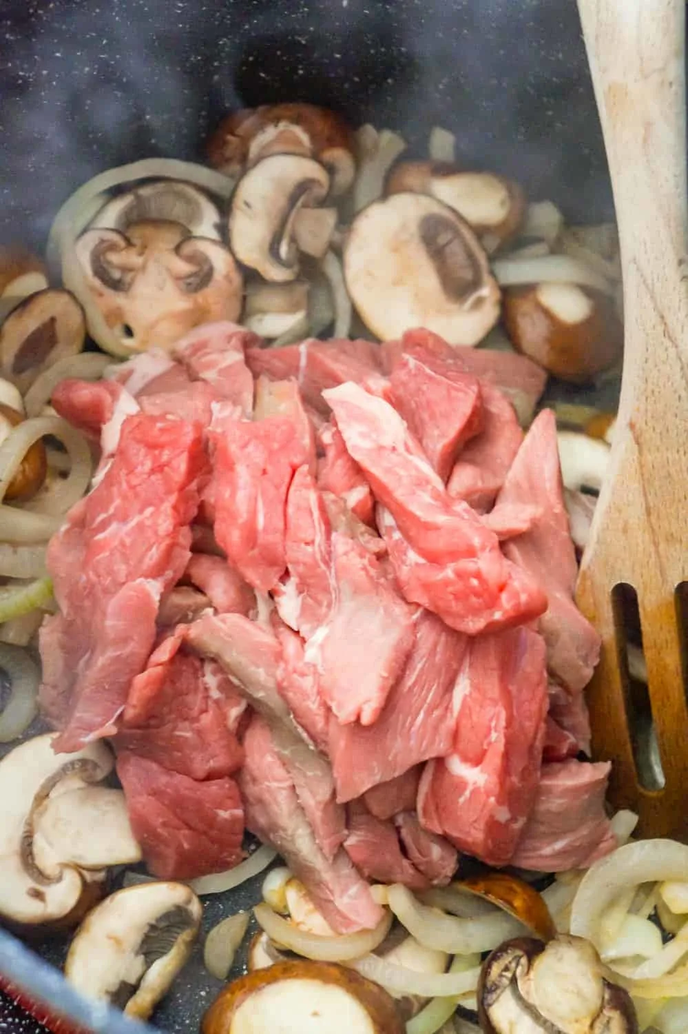 raw steak strips on top of sliced mushrooms and onions in a large pan