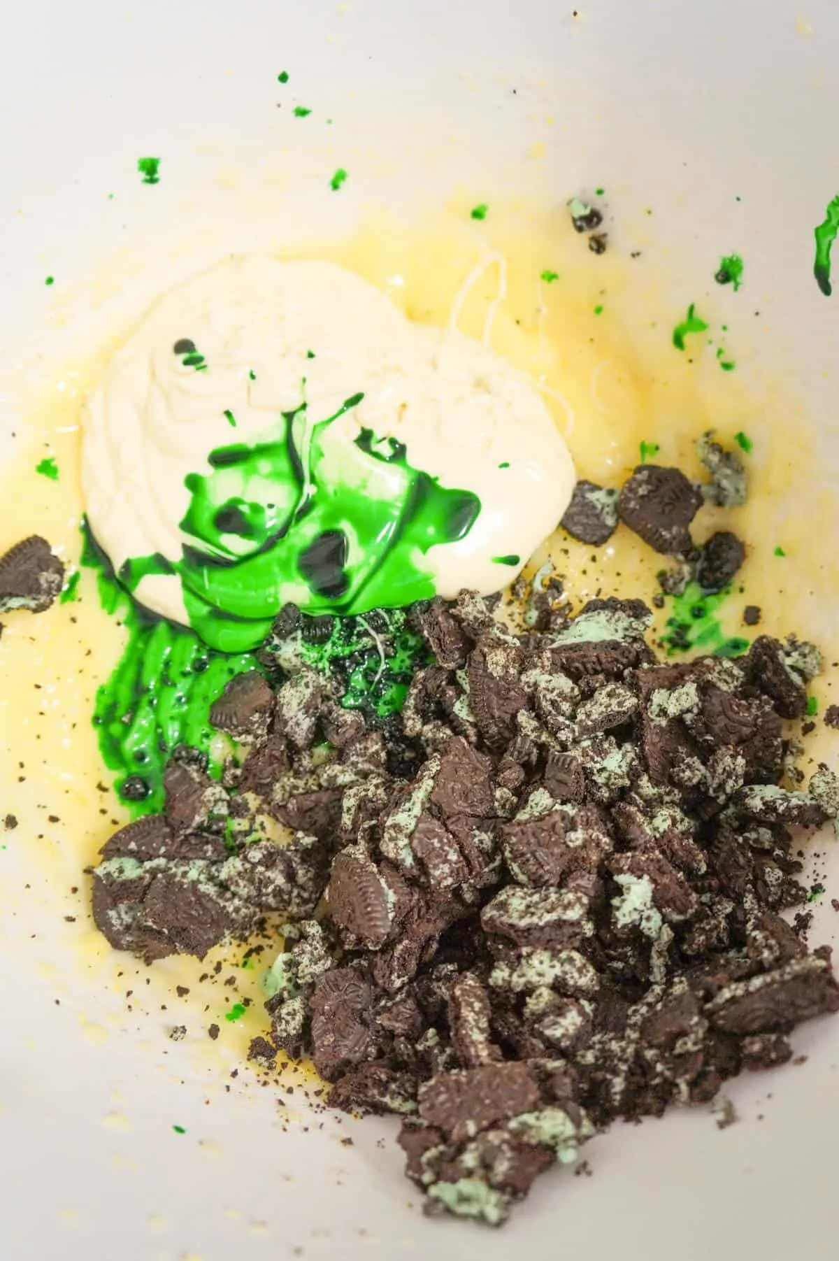 green food colouring, melted white chocolate and crumbled mint oreo cookies on top of vanilla pudding mixture in a mixing bowl