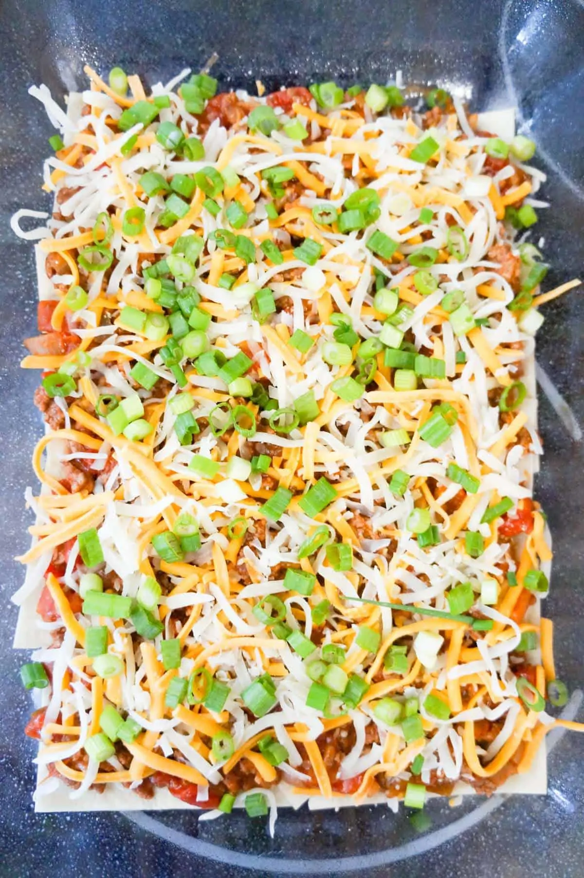chopped green onions and shredded cheese on top of meat and tortillas in a baking dish