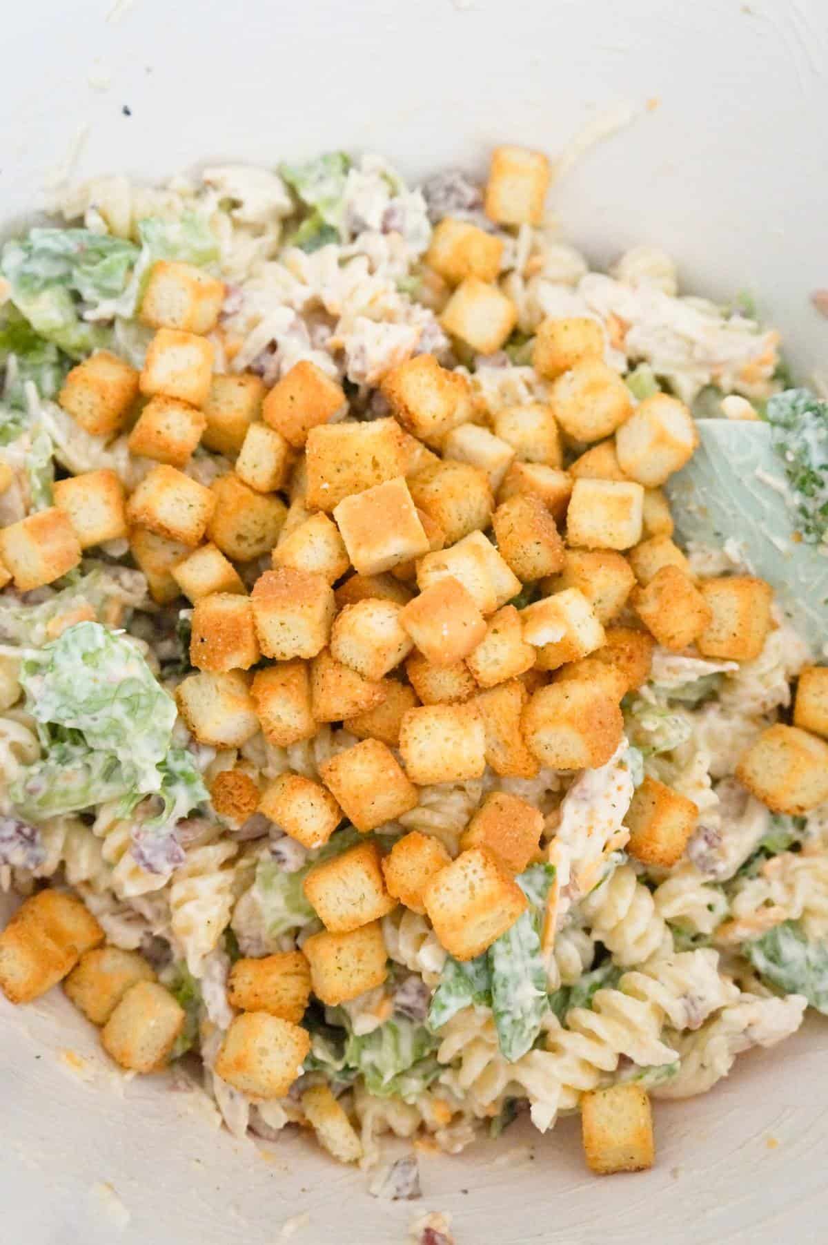 croutons on top of chicken caesar pasta salad in a mixing bowl