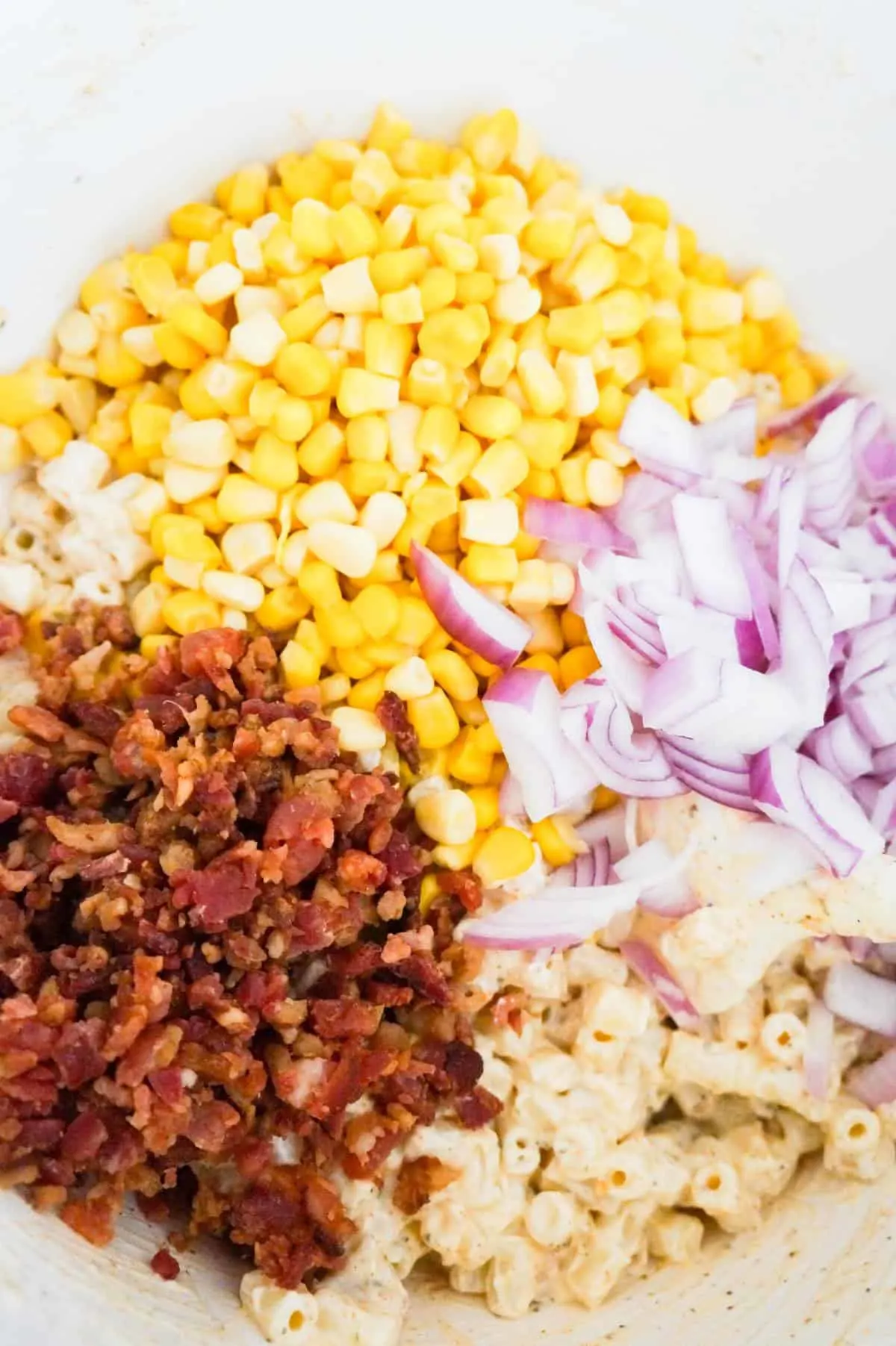 crumbled bacon, canned corn and diced red onions on top of macaroni salad in a mixing bowl