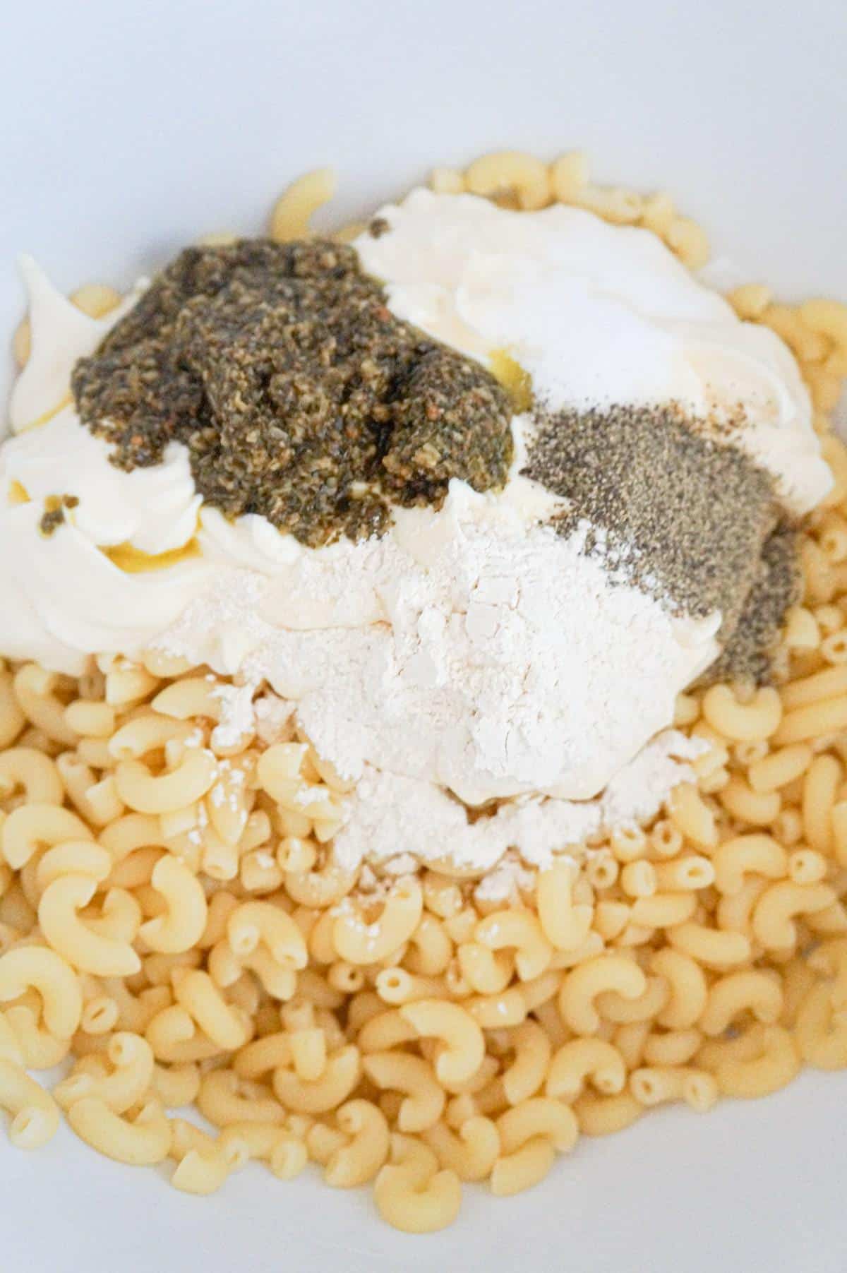 basil pesto, mayo and spices on top of cooked macaroni noodles in a mixing bowl