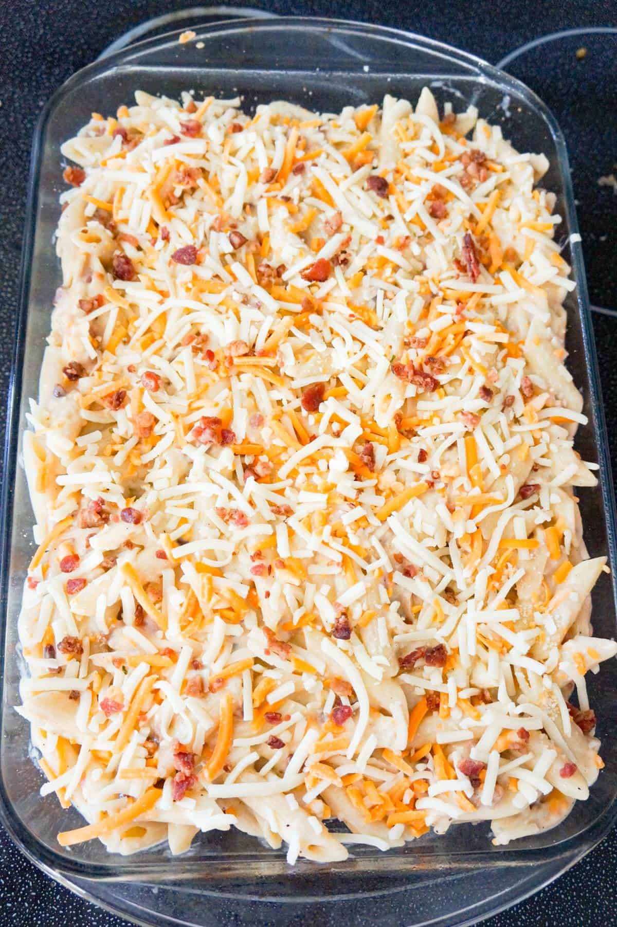 crumbled bacon and shredded cheddar on top of penne pasta in a baking dish
