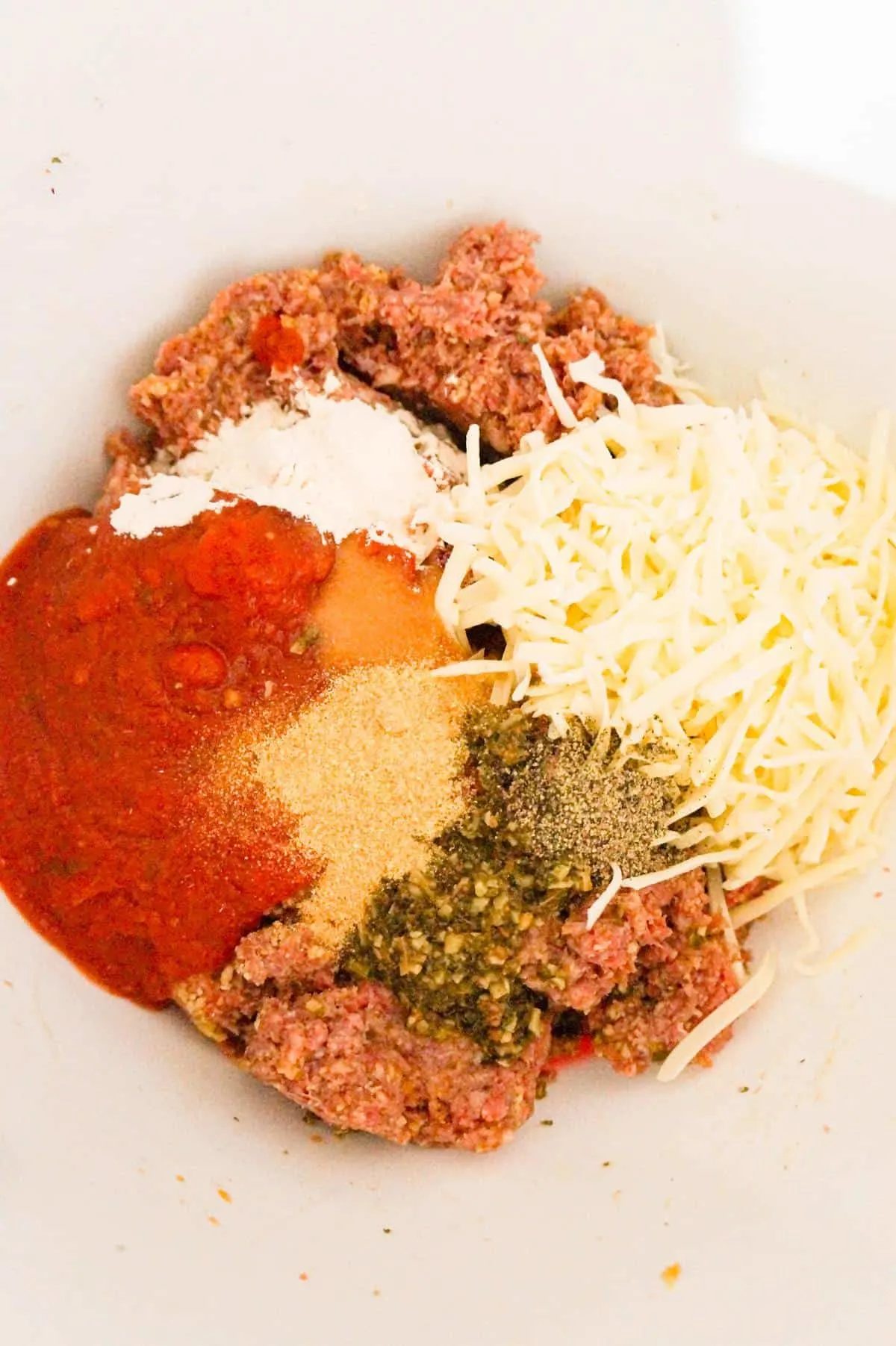 spices, shredded cheese and marinara sauce on top of ground beef mixture in a mixing bowl