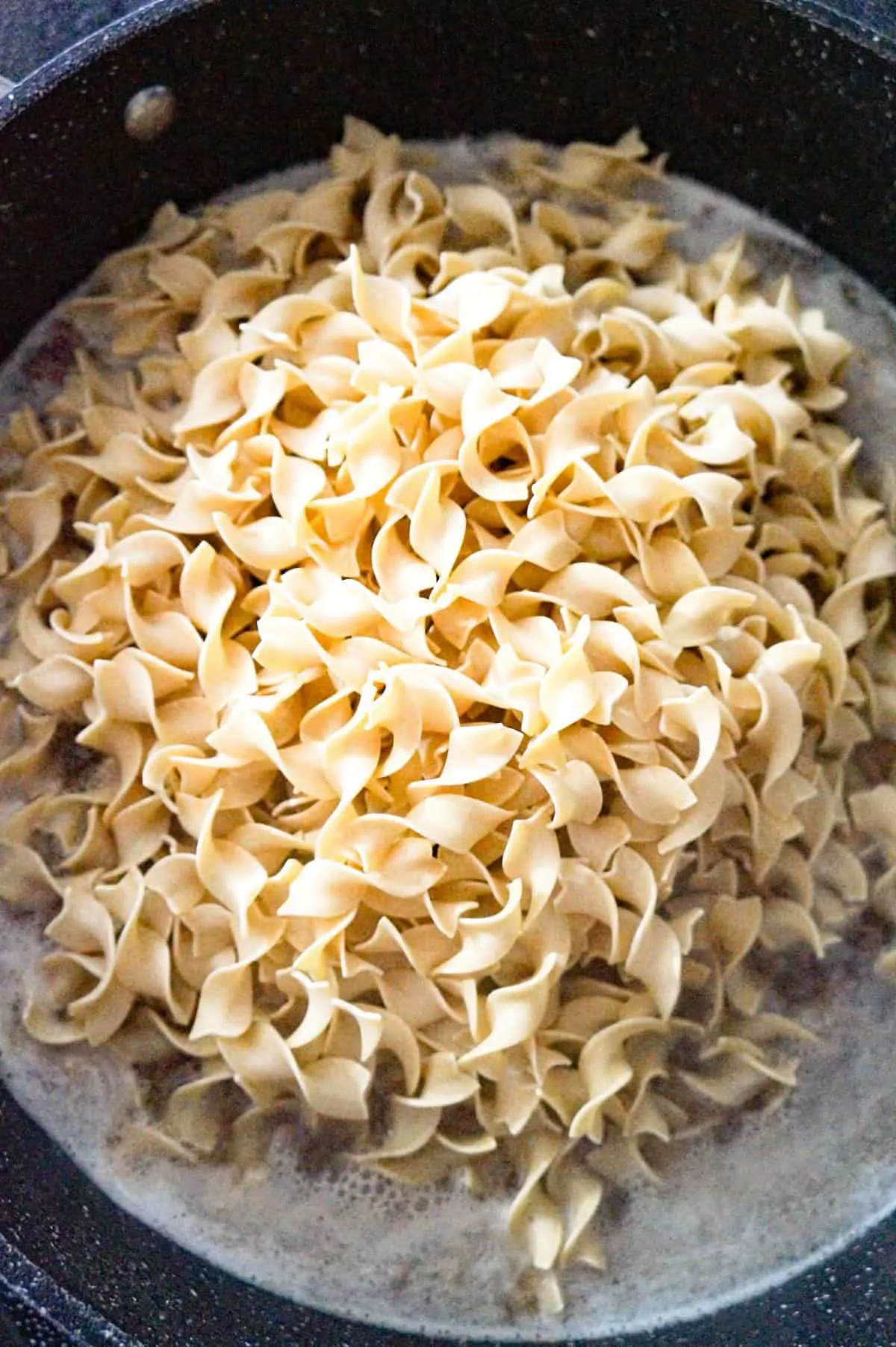 uncooked egg noodles on top of cooked ground beef and boiling water in a pan