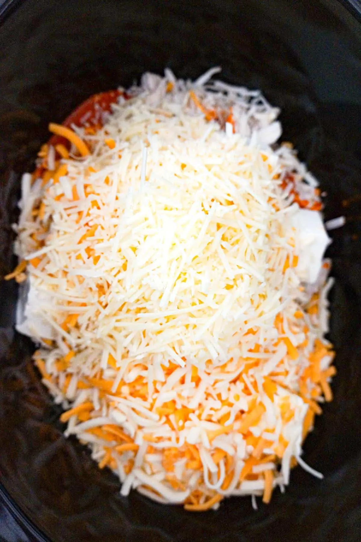 shredded parmesan and shredded Mexican cheeses on top of buffalo chicken dip ingredients in a crock pot