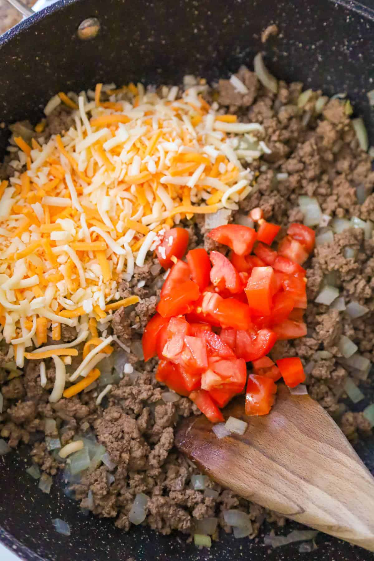 shredded cheddar cheese and diced tomatoes on top of cooked ground beef in a saute pan