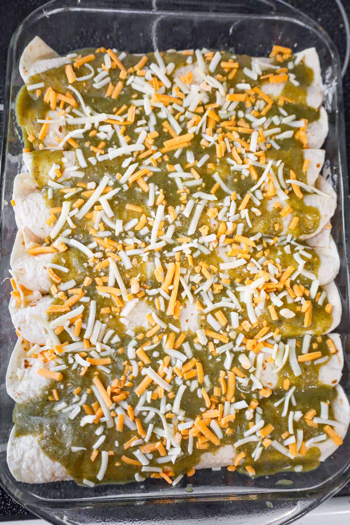 shredded cheese and green enchilada sauce on top of rolled tortillas in a baking dish