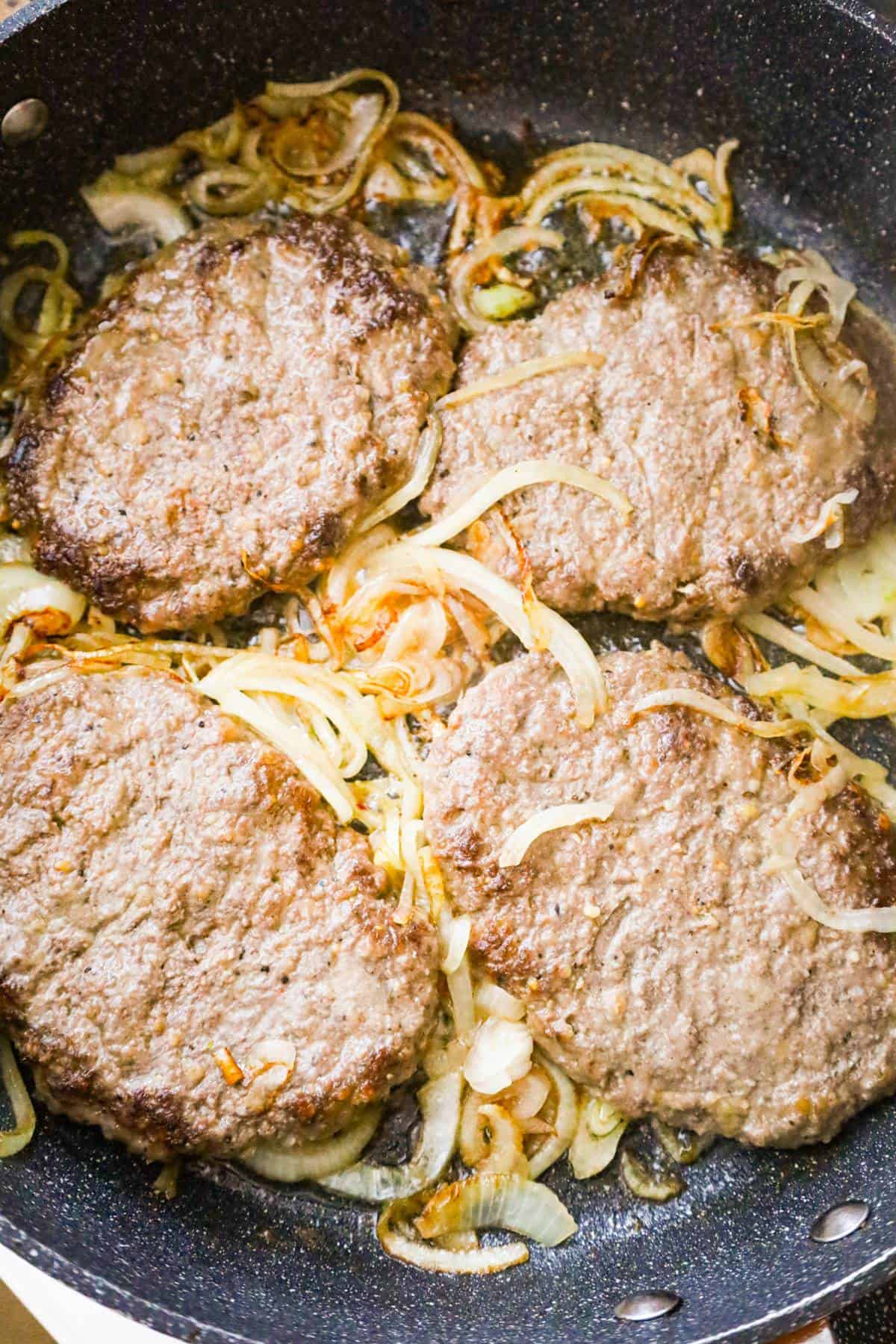 hamburger patties cooking on top of sliced onions in a pan