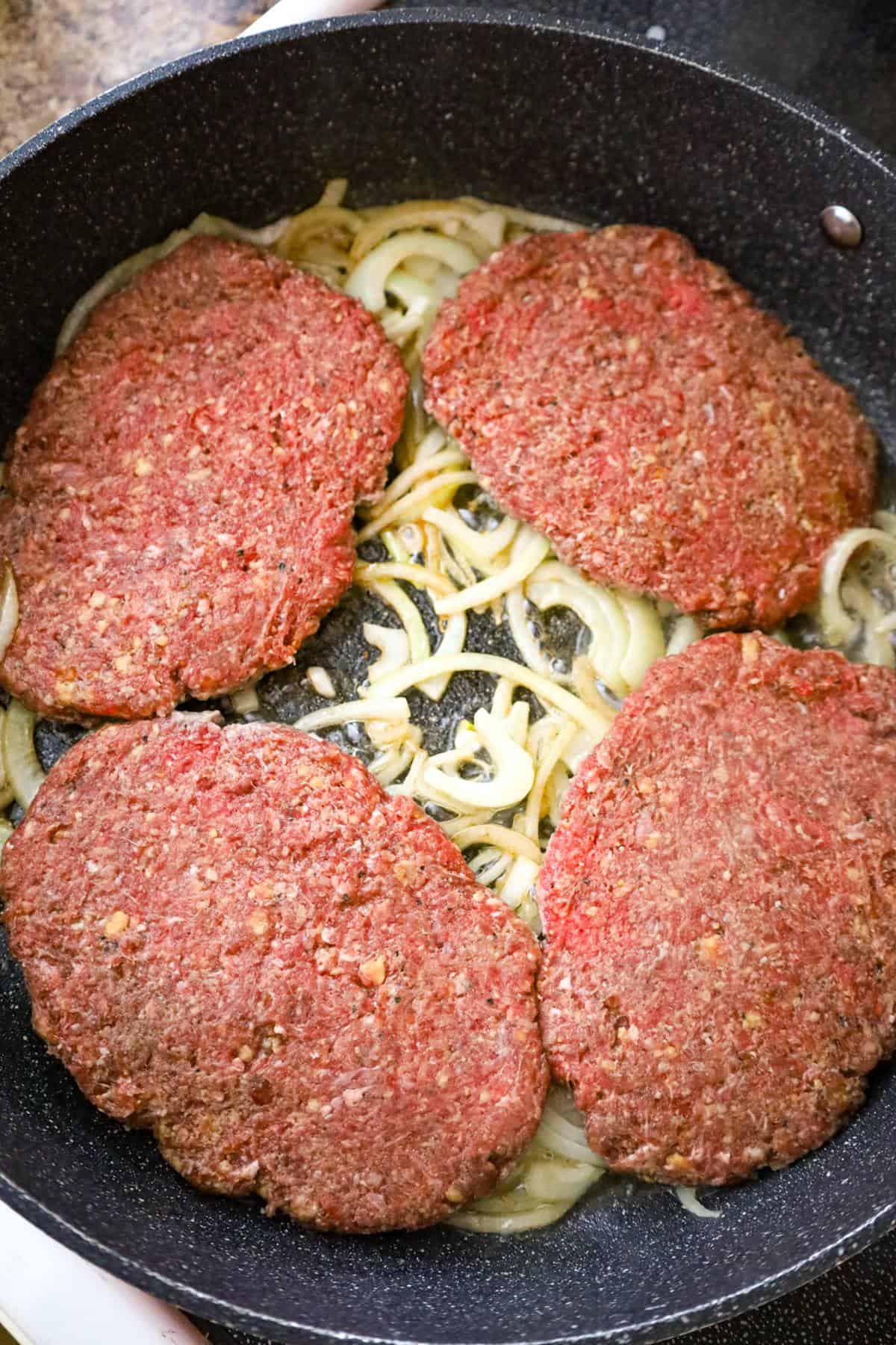 raw hamburger patties on top of sliced onions in a pan