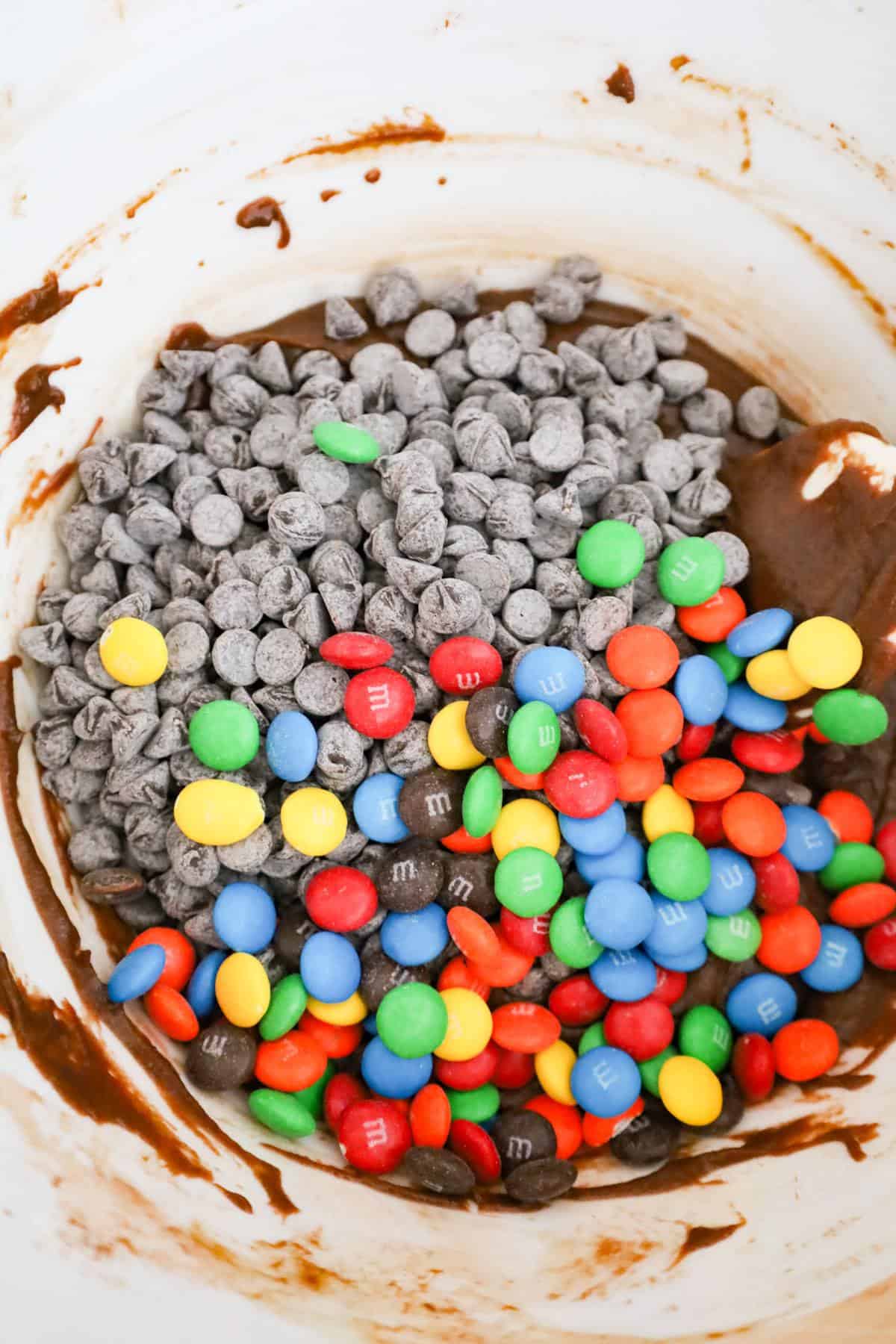 semi sweet chocolate chips and M&Ms on top of brownie batter in a mixing bowl