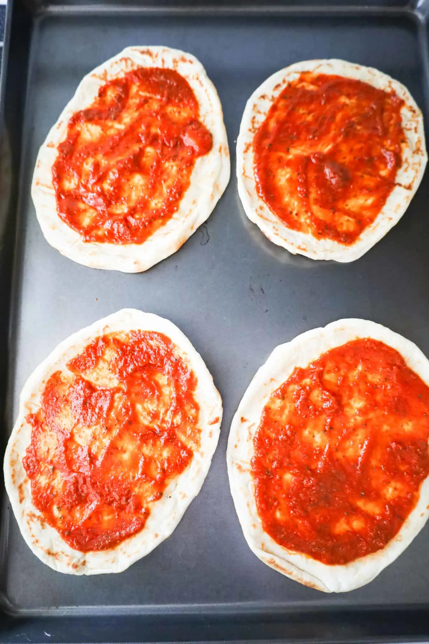 pizza sauce spread on naan breads on a baking sheet