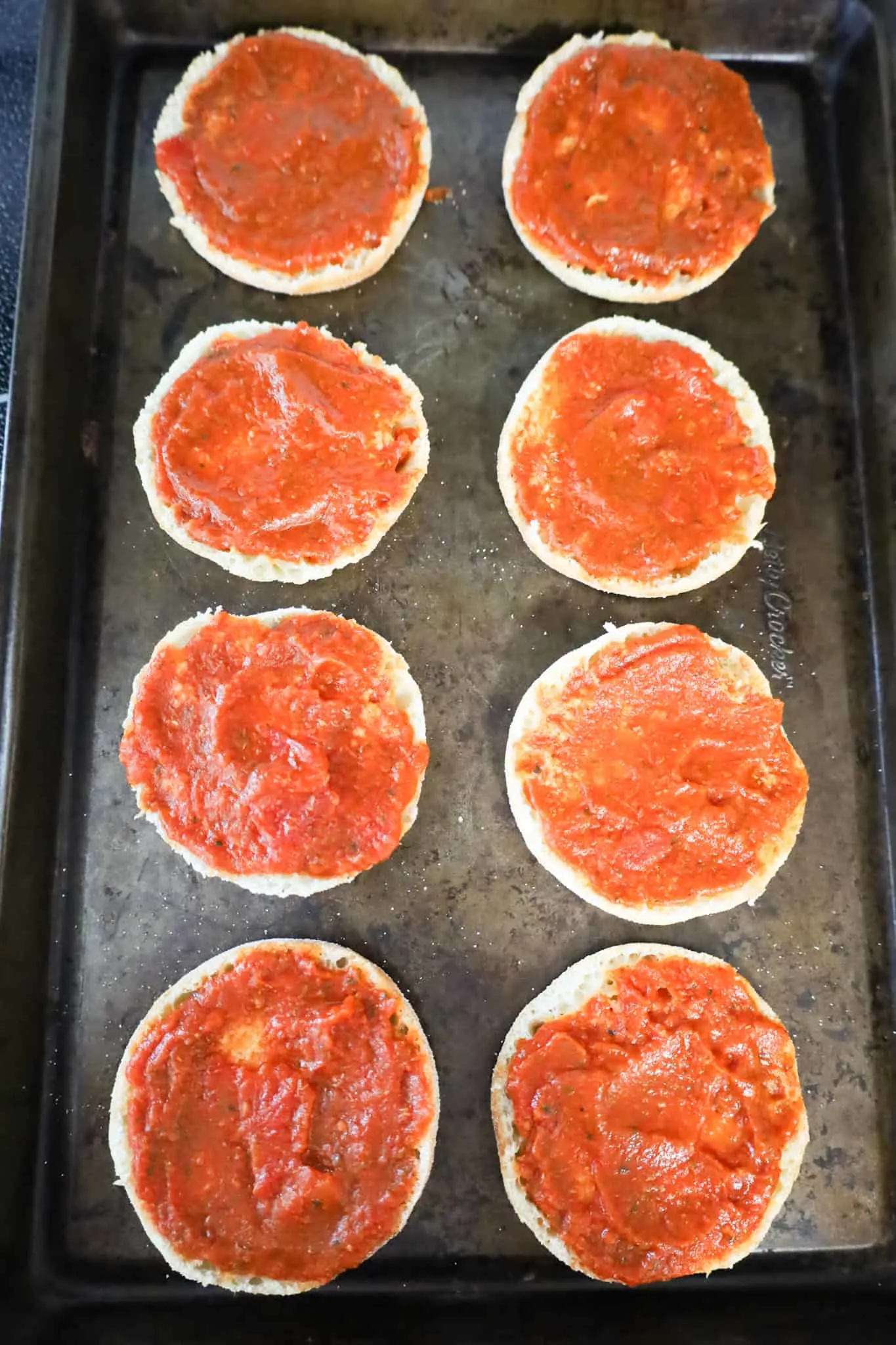 pizza sauce spread on top of split English muffins on a baking sheet