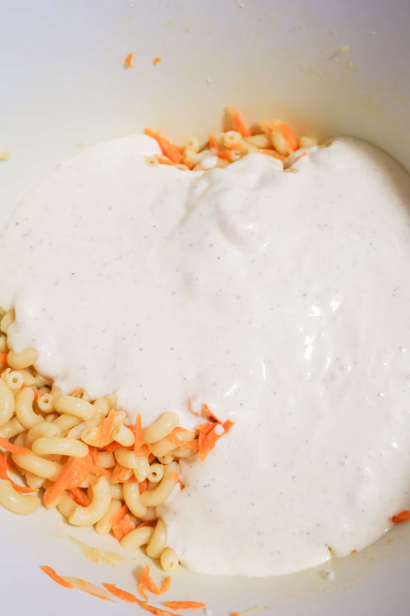 creamy mayo dressing poured over macaroni and grated carrot in a mixing bowl