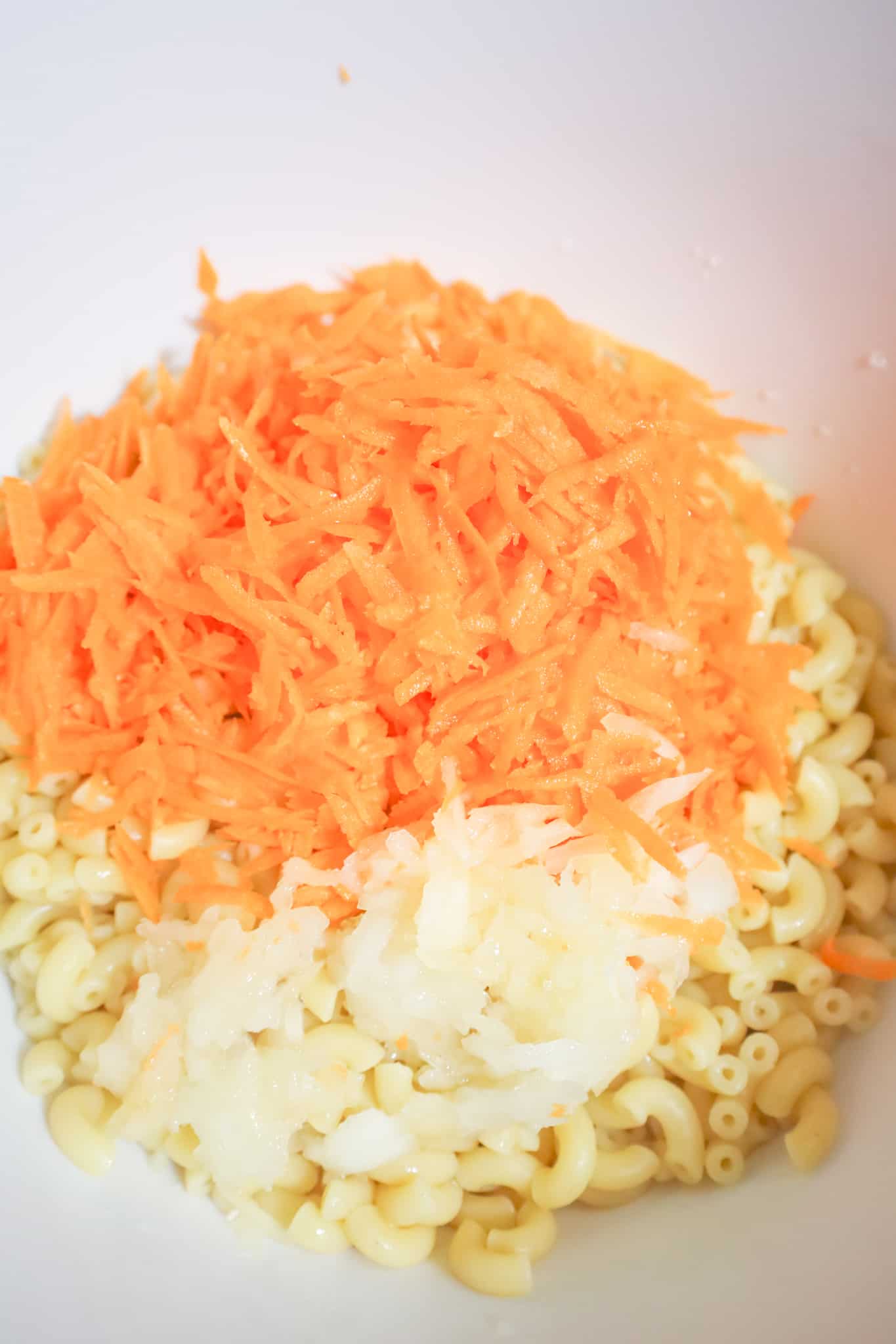 grated carrot and grated onion on top of cooked macaroni in a mixing bowl