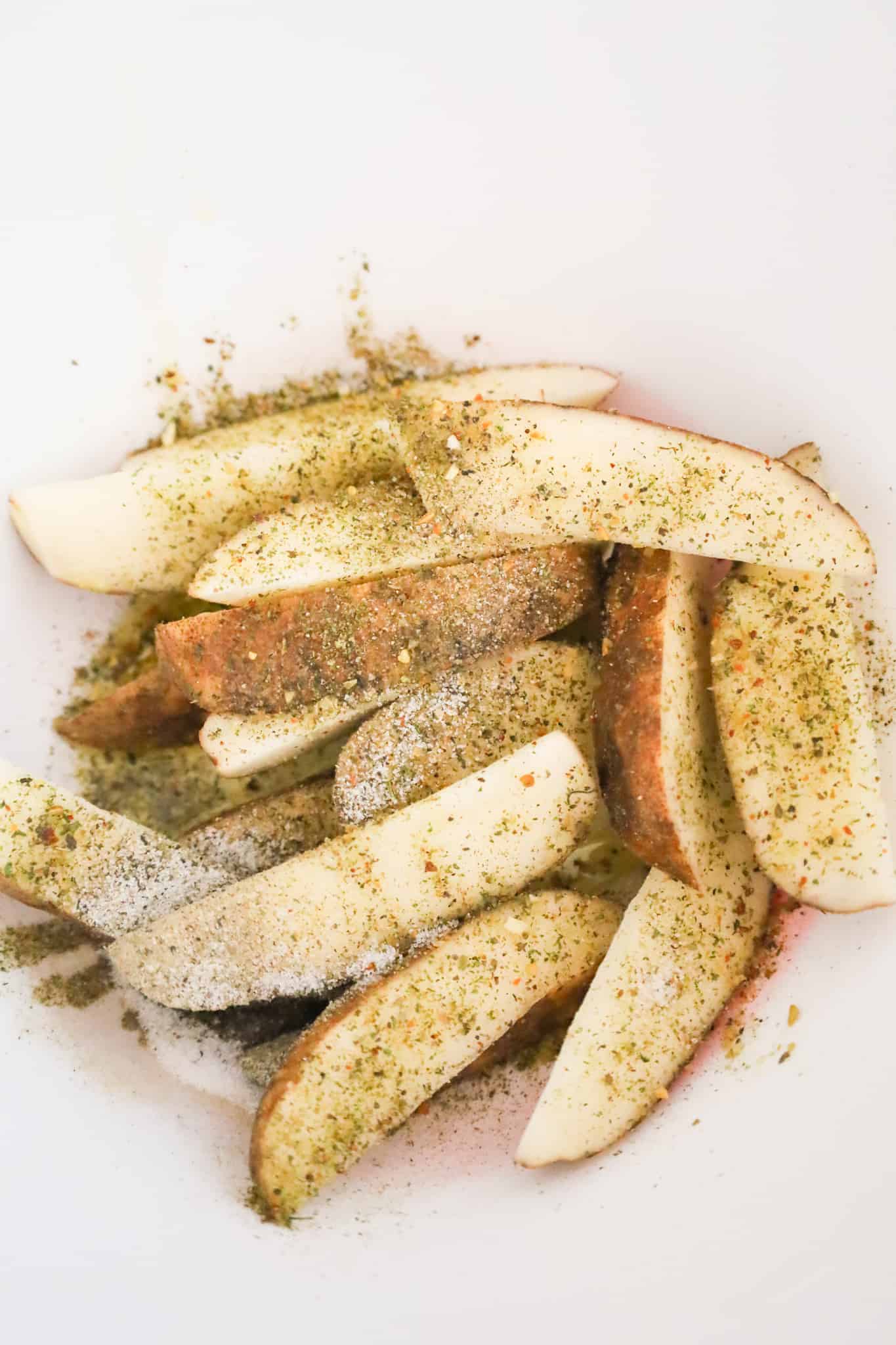 olive oil and spices on top of raw potato wedges in a mixing bfowl