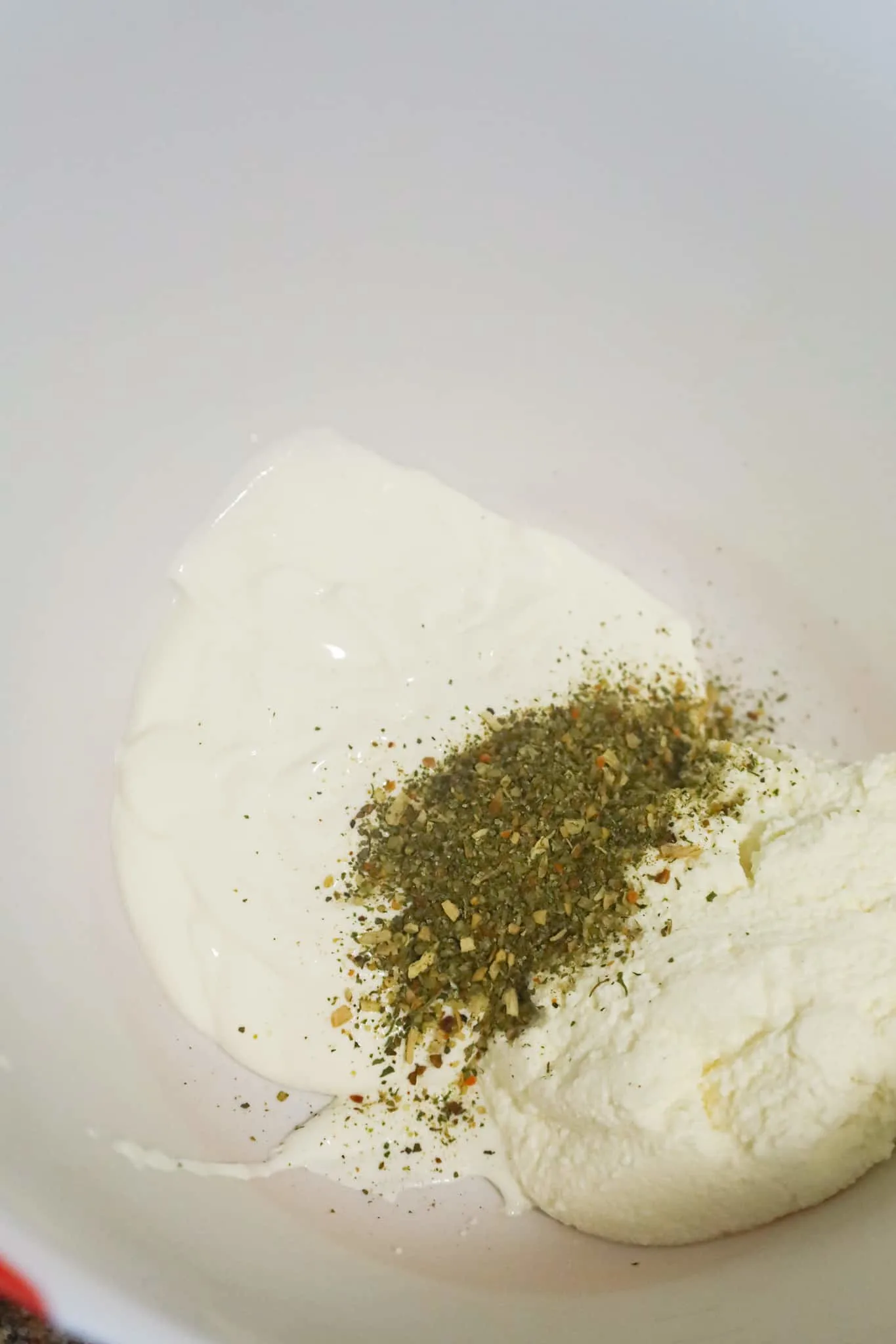 Italian seasoning on top of sour cream and ricotta in a mixing bowl