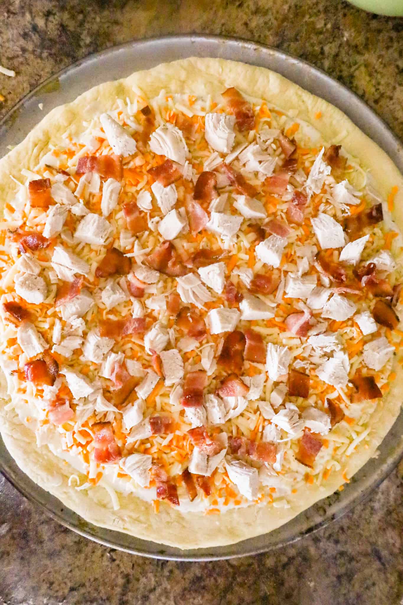 chopped bacon and chicken on top of shredded mozzarella and cheddar cheese on top of pizza dough before baking