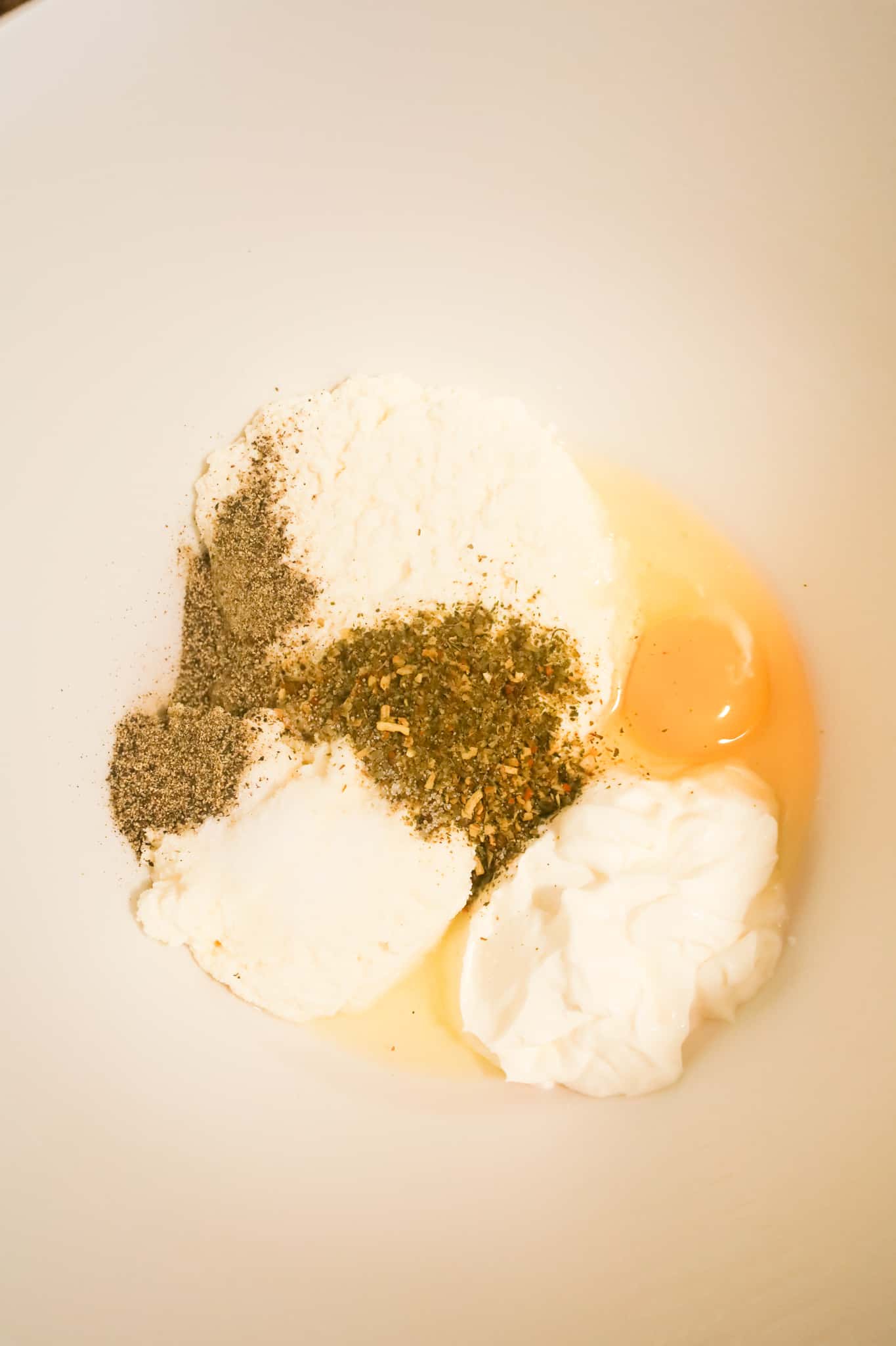 spices on top of ricotta, sour cream and an egg in a mixing bowl