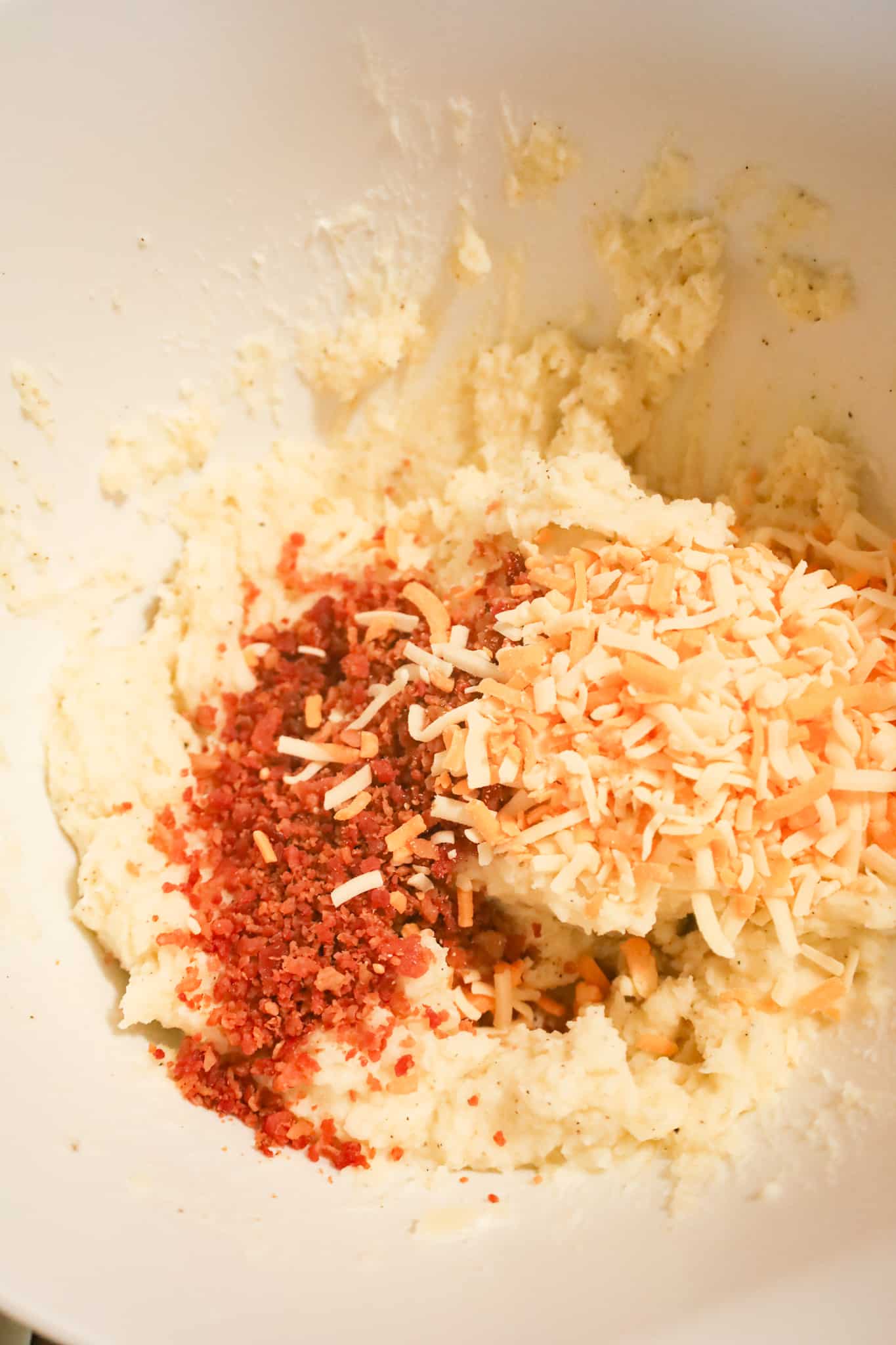 crumbled bacon and shredded cheese on top of mashed potato mixture in a mixing bowl
