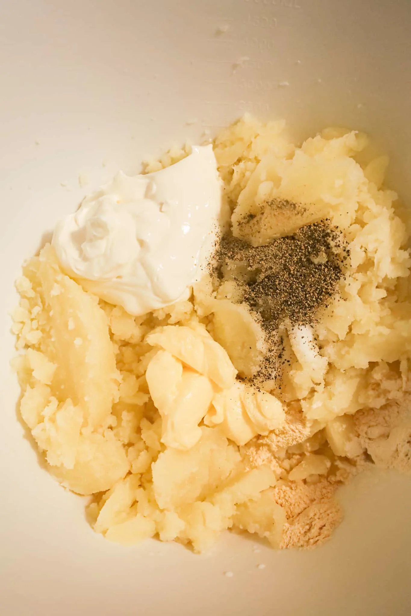 butter, sour cream and spices on top of mashed potatoes in a mixing bowl