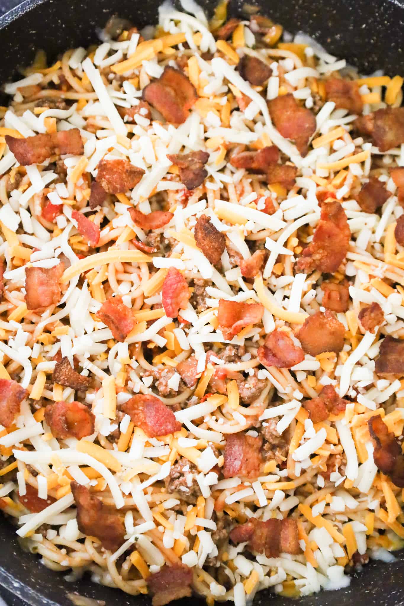 shredded cheese and chopped bacon on top of spaghetti in a skillet