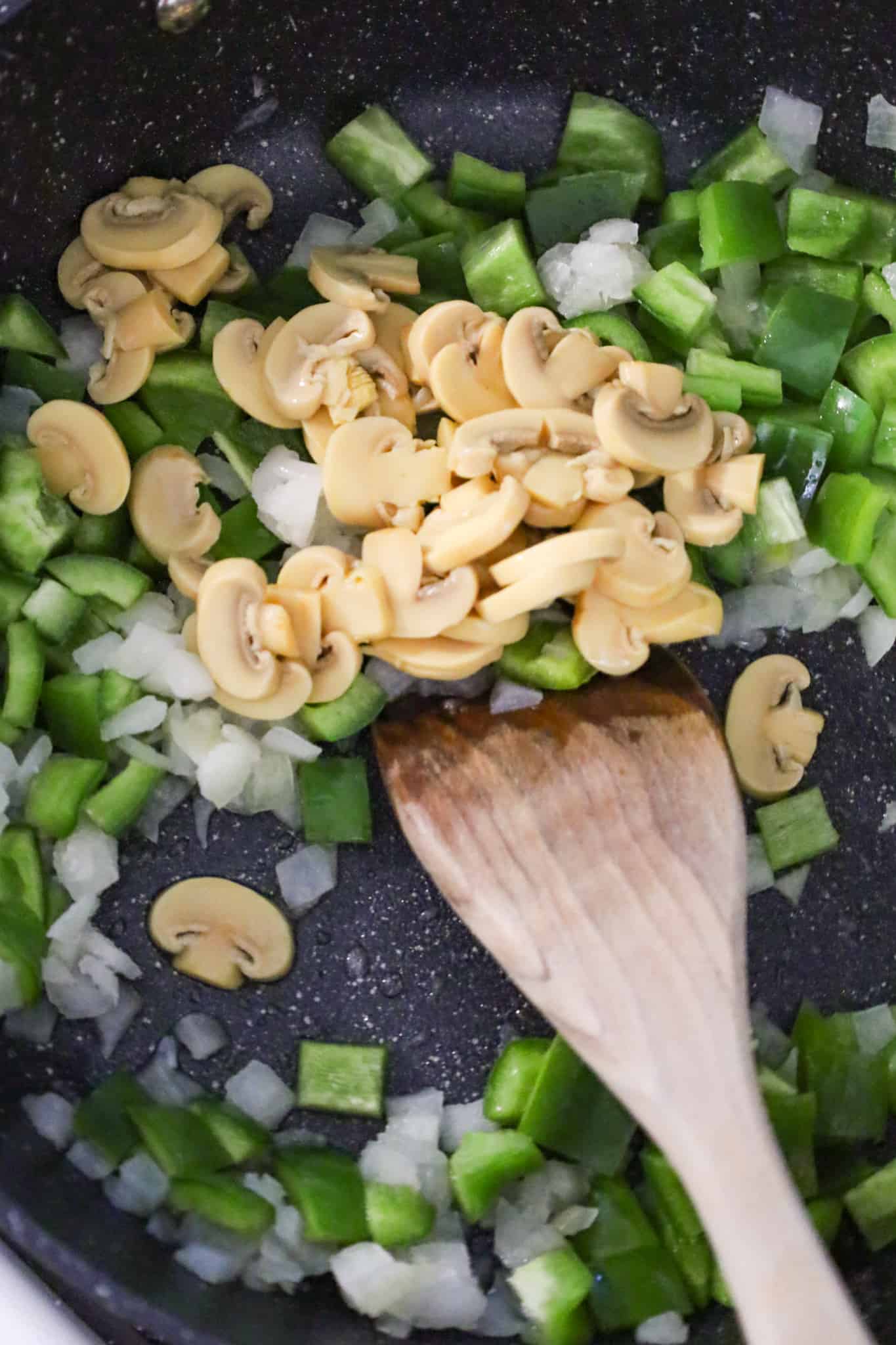 sliced mushrooms on top of diced onions and diced green peppers in a skillet