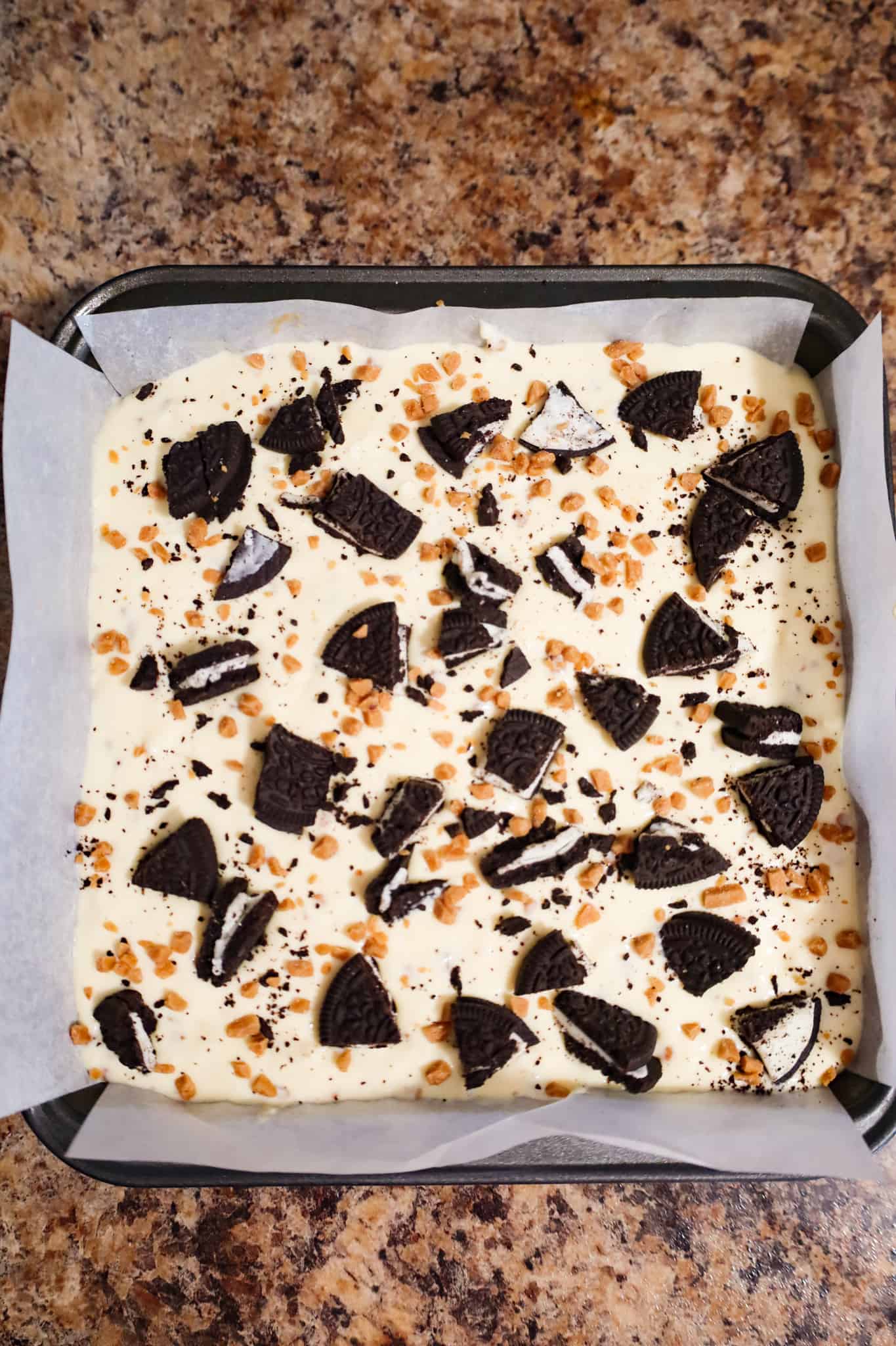 crumbled oreos and toffee bits on top of cheesecake mixture in a baking pan