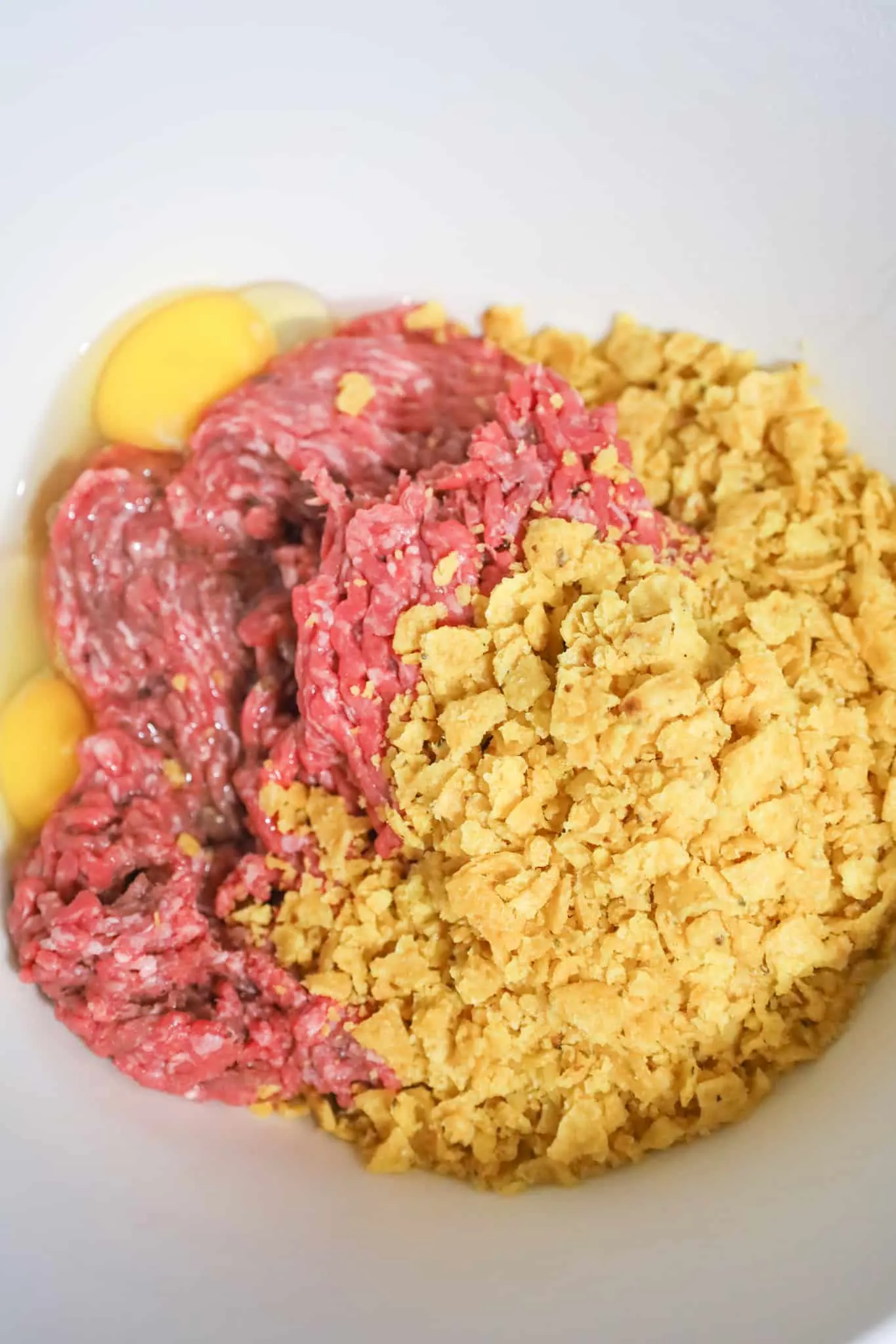 crushed corn chips and eggs on top of raw ground beef in a mixing bowl