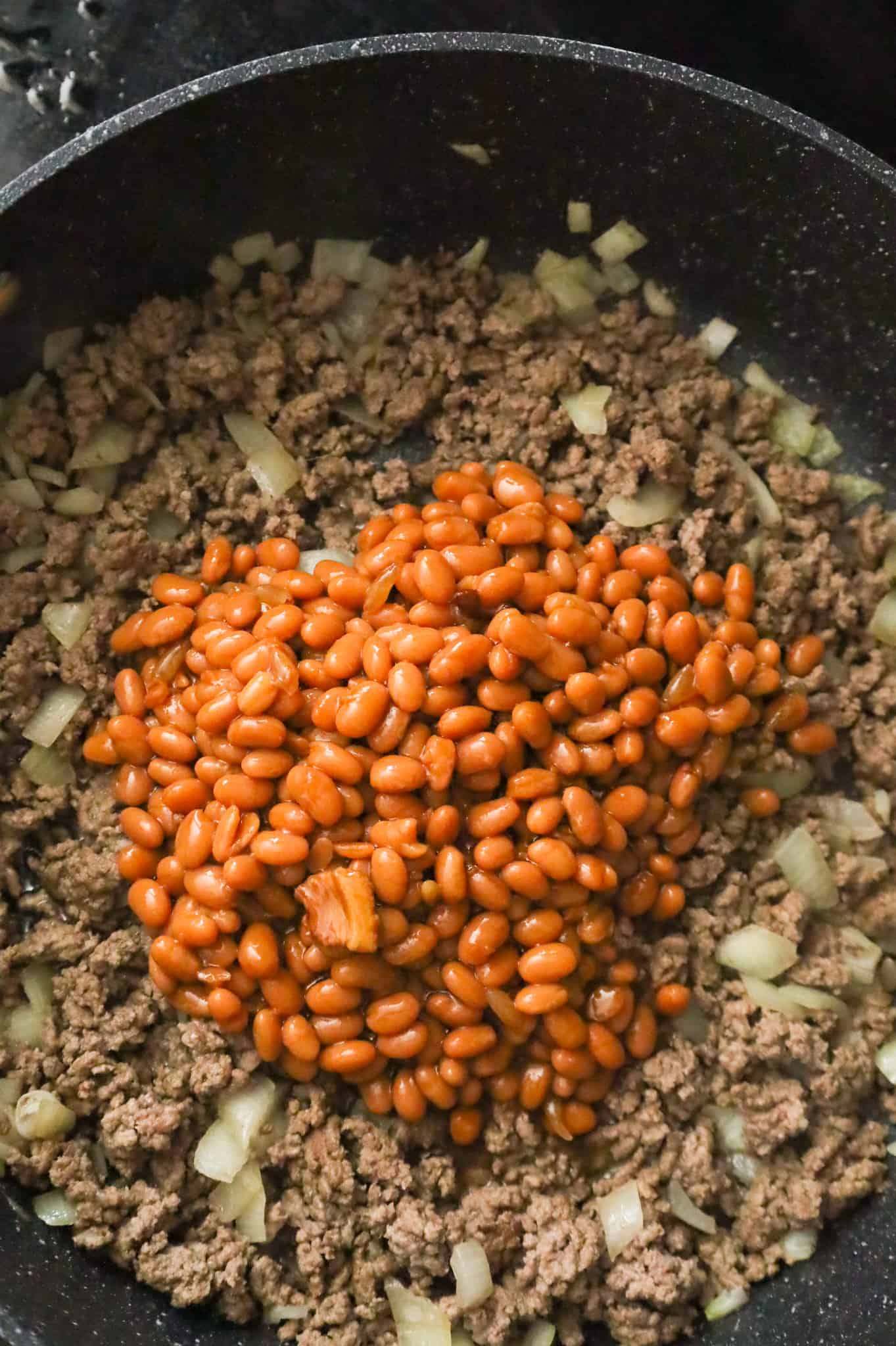 baked beans on top of cooked ground beef and diced onions in a skillet