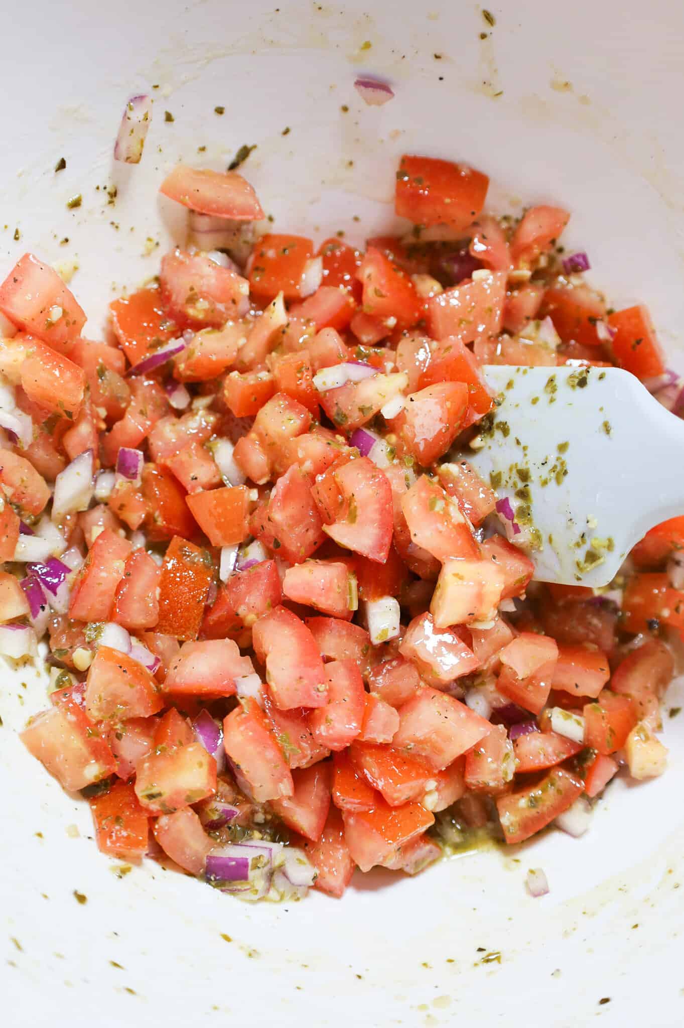 diced tomato, red onion, basil pesto and olive oil mixture being stirred in a mixing bowl