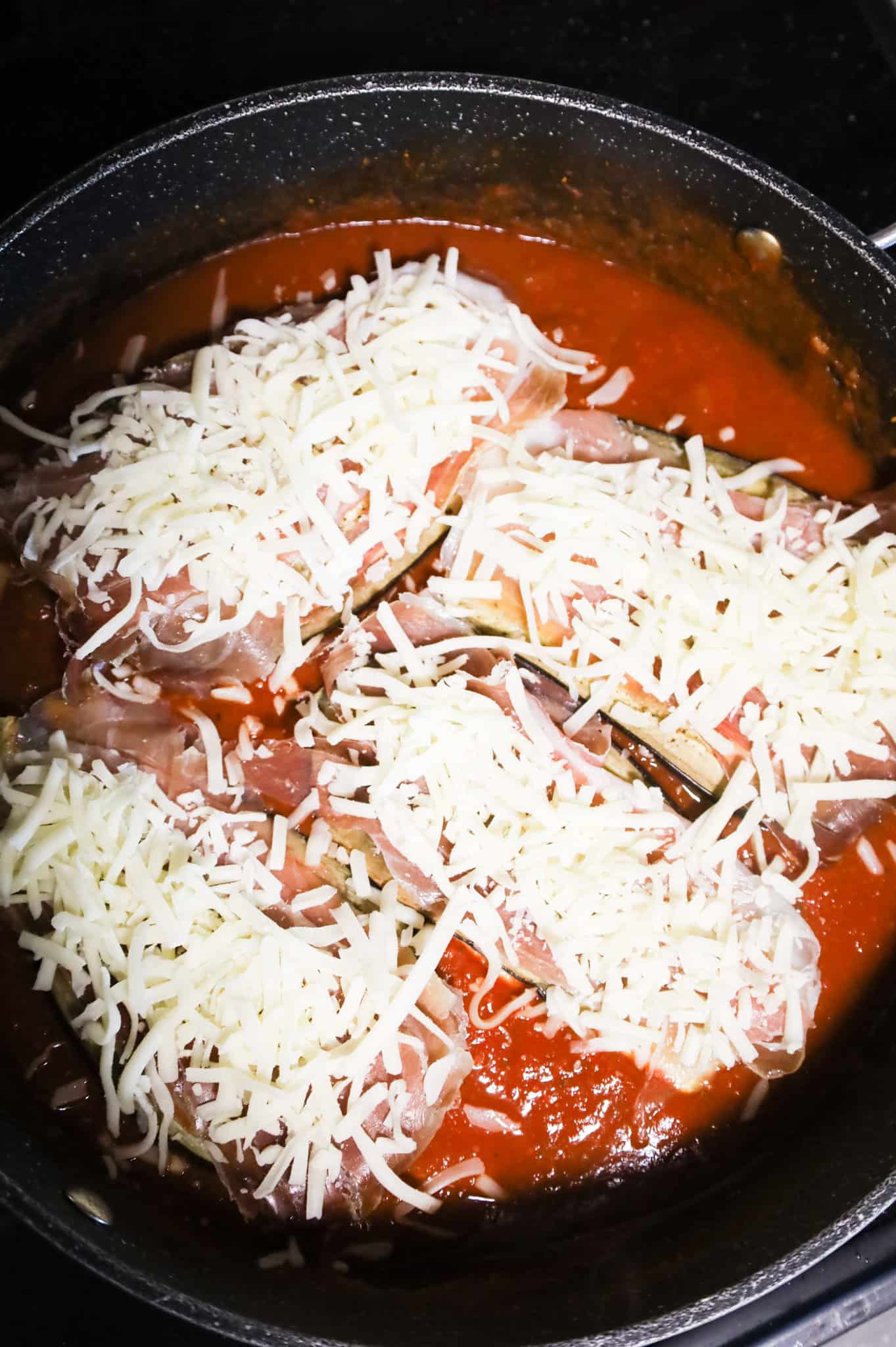 shredded cheese on top of prosciutto, eggplant and chicken cutlets in a skillet with tomato sauce