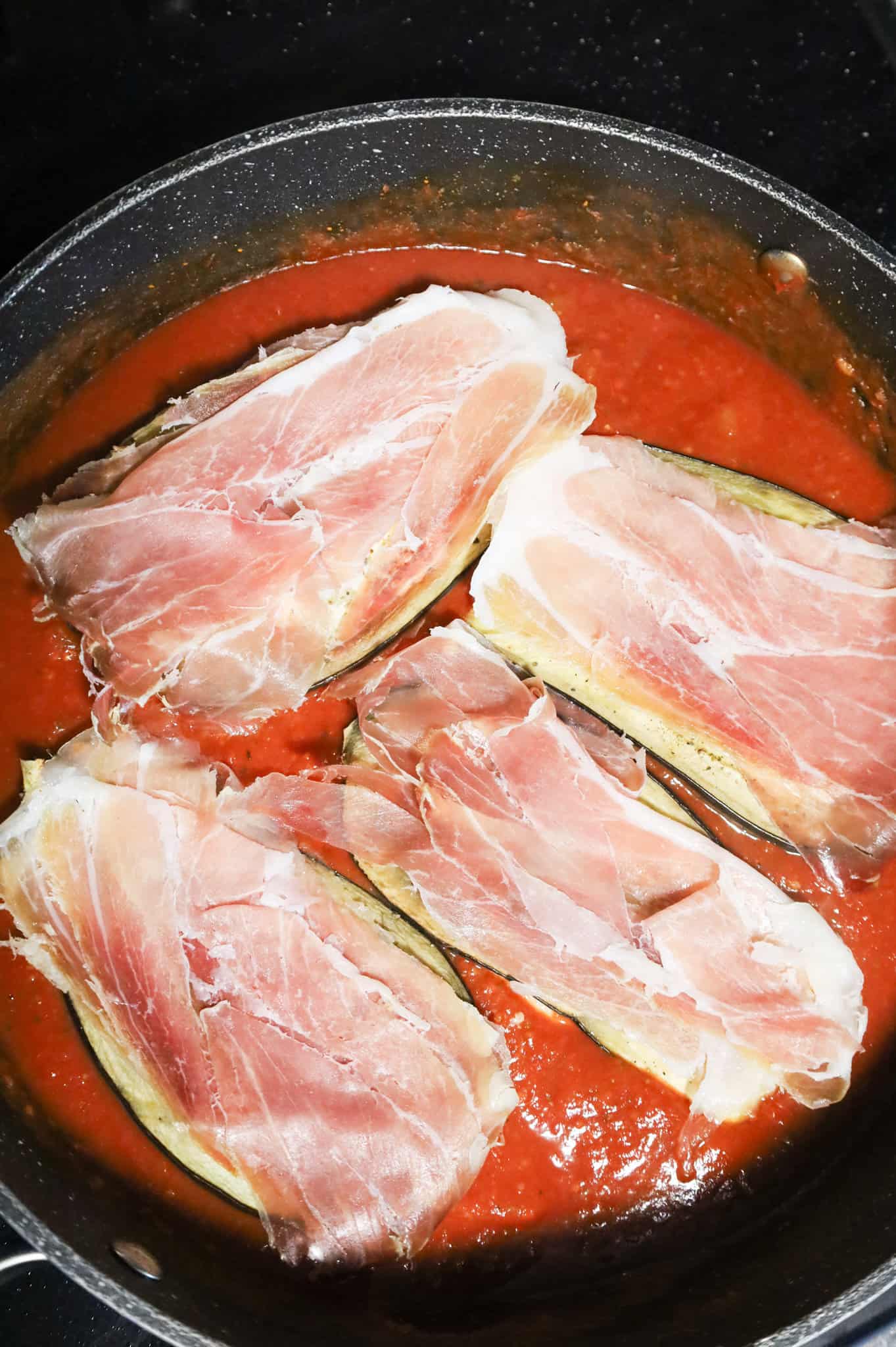 prosciutto and eggplant on top of chicken cutlets in a skillet with tomato sauce