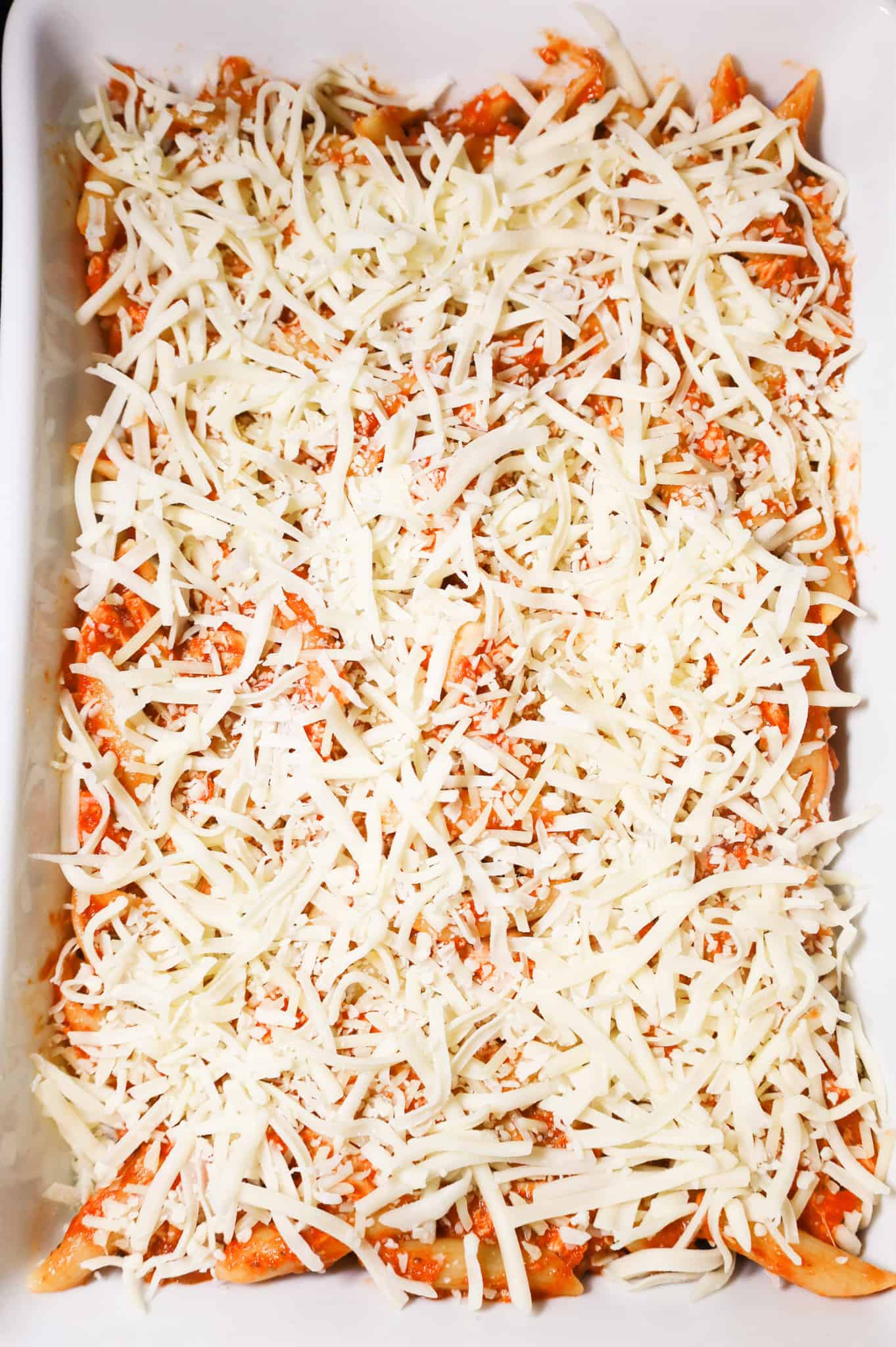 shredded mozzarella sprinkled on top of penne marinara in a baking dish