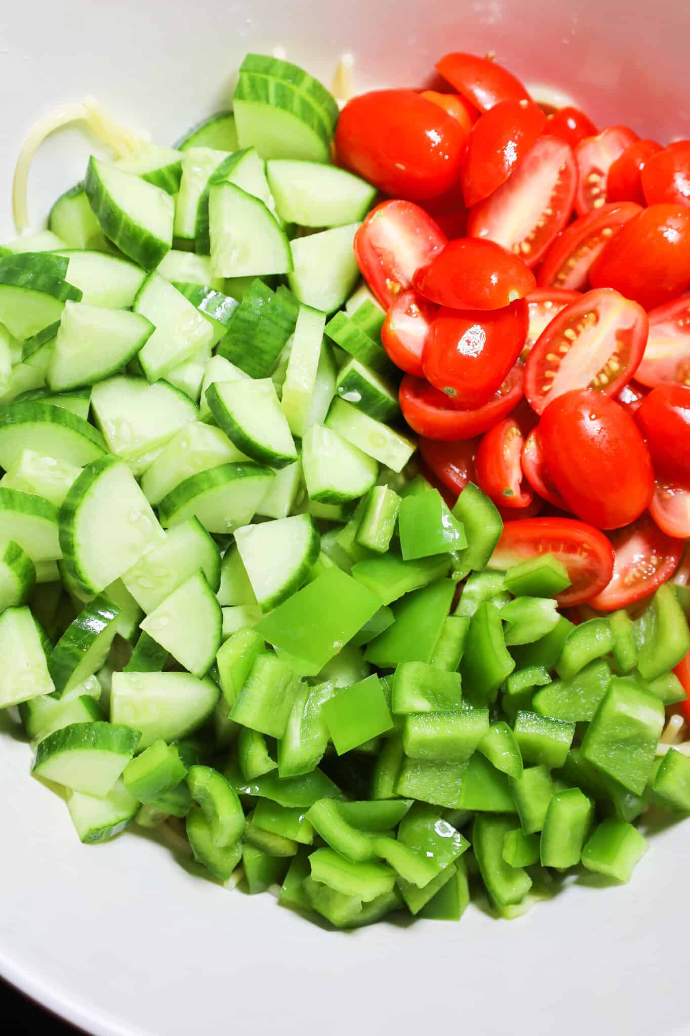 diced cucumbers, diced green peppers and halved grape tomatoes on top of cooked spaghetti in a mixing bowl