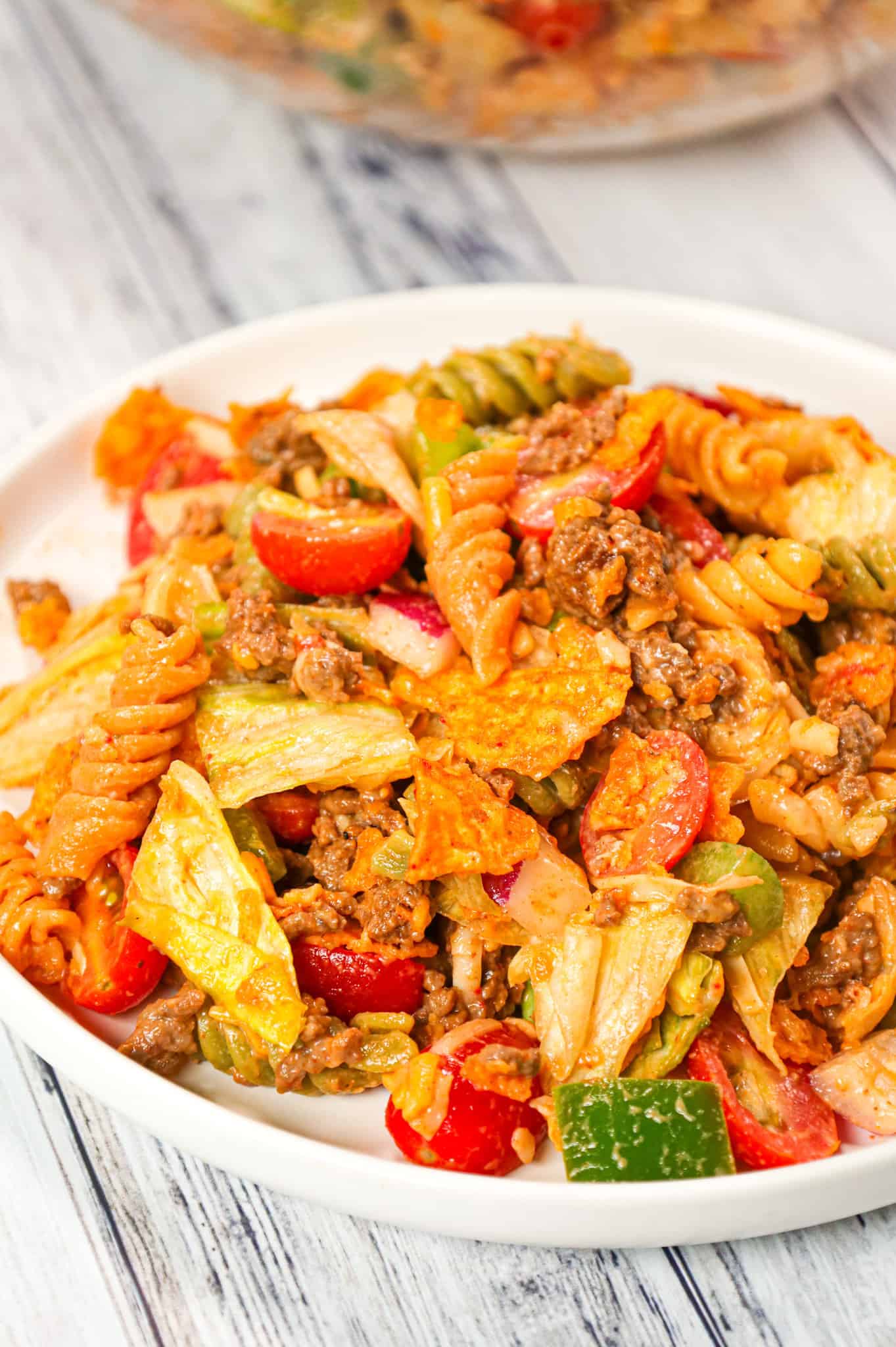 Taco Pasta Salad is the perfect party side dish loaded with tri-colour rotini pasta, taco seasoned ground beef, iceberg lettuce, onions, tomatoes, green peppers, shredded cheese, salsa, ranch dressing and crumbled Doritos nacho chips.