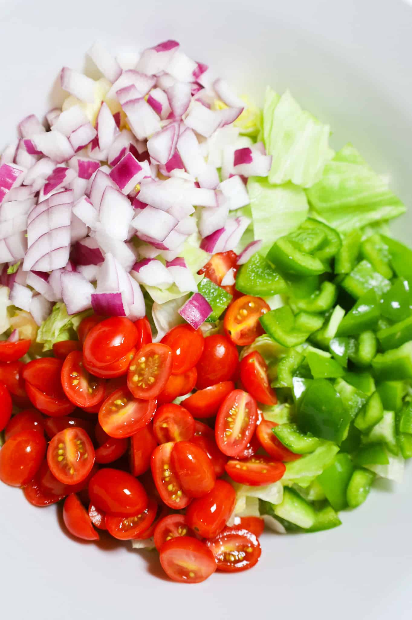 halved grape tomatoes and diced red onions and green peppers on top of iceberg lettuce in a mixing bowl