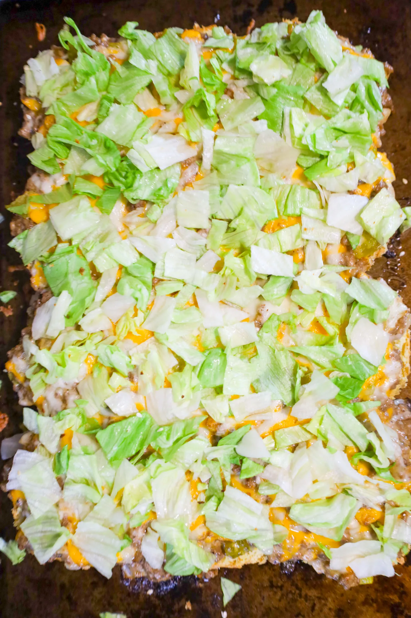 shredded iceberg lettuce on top of cheesy ground beef buns on a baking sheet