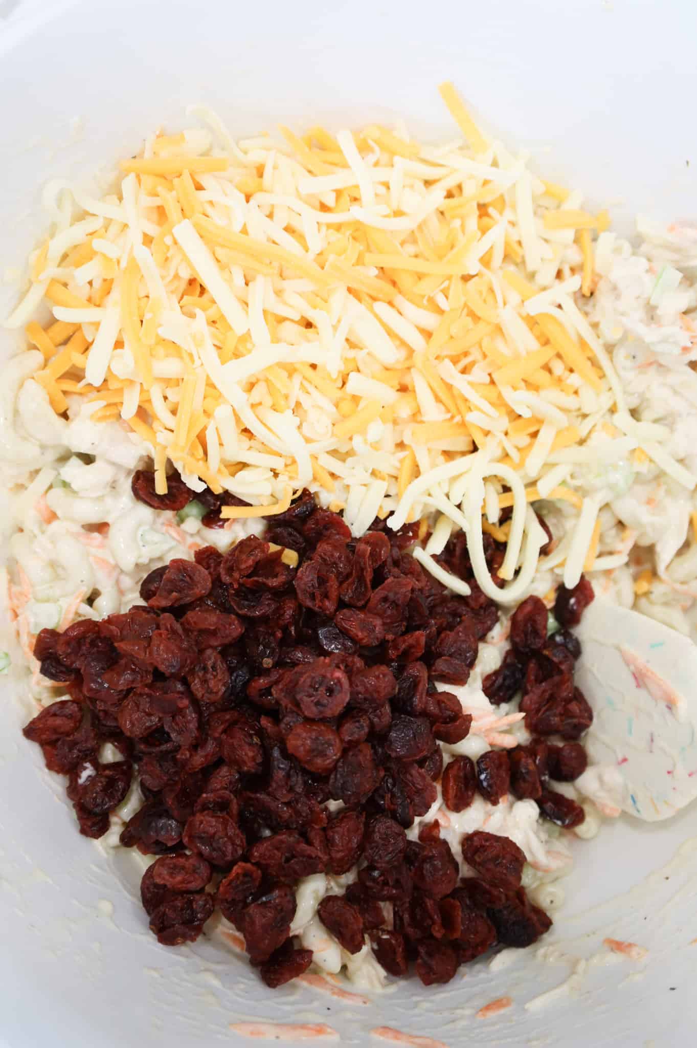 shredded cheddar and dried cranberries on top of macaroni salad in a mixing bowl