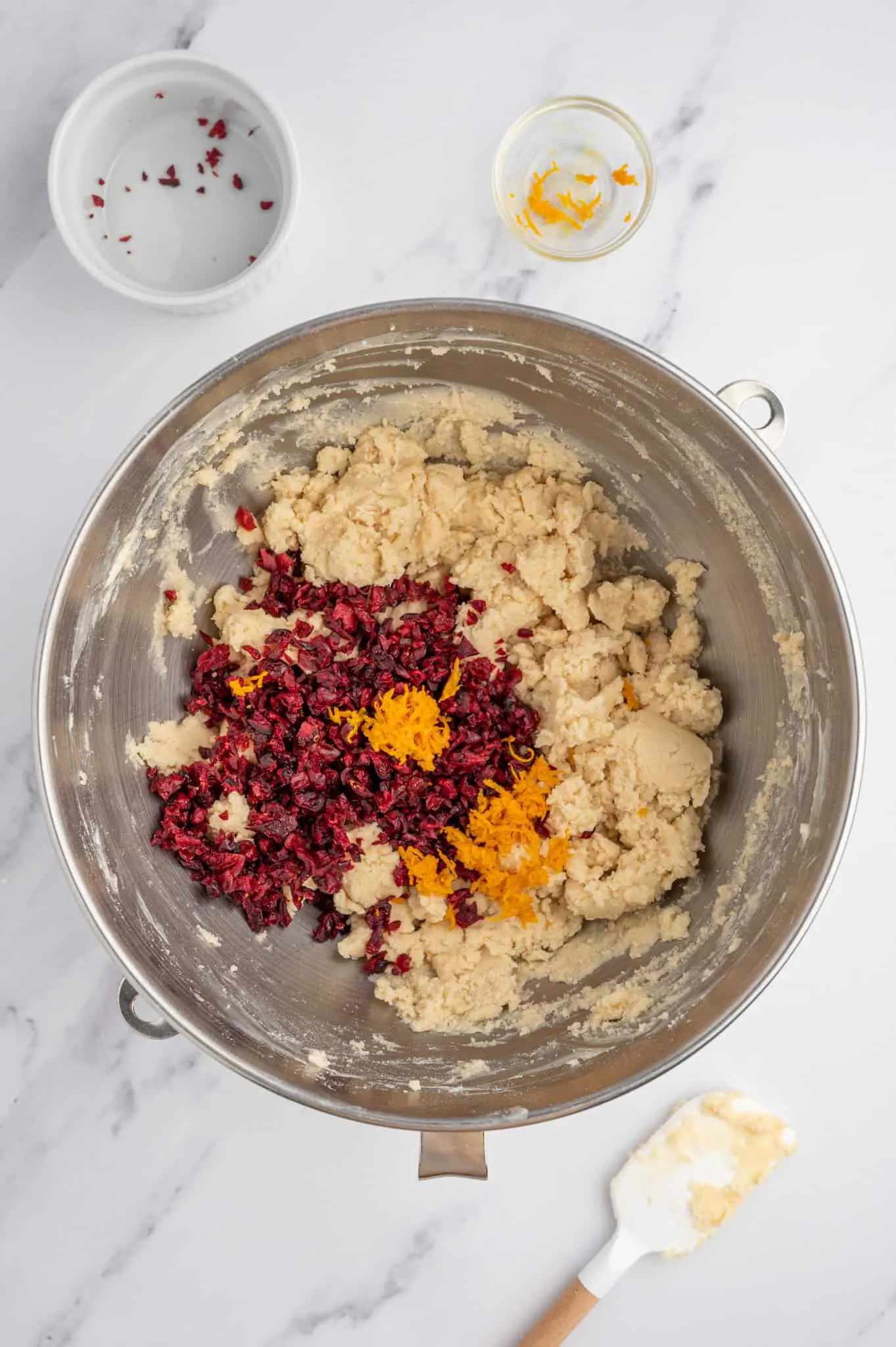 orange zest and dried cranberries on top of shortbread cookie dough in a mixing bowl