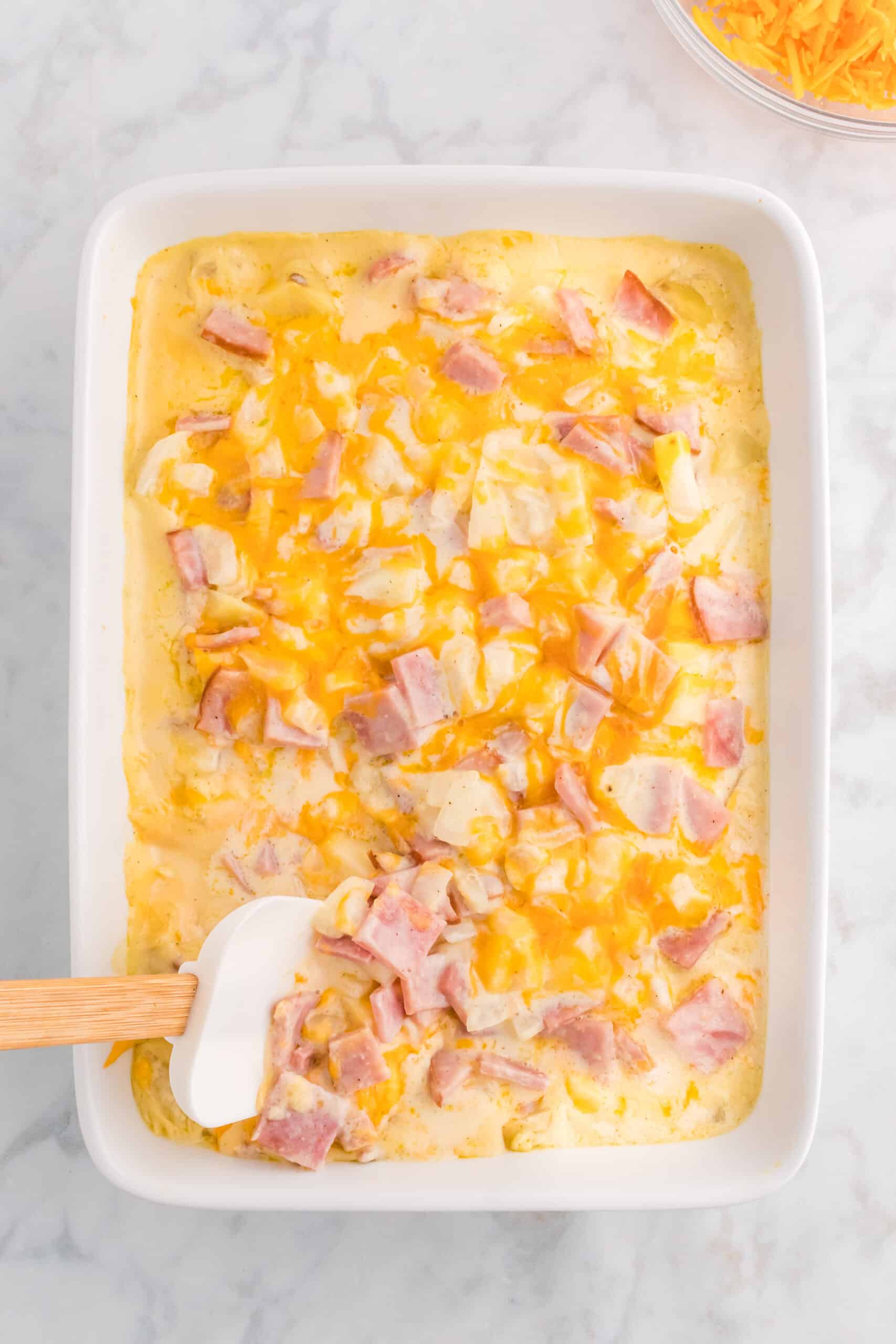 ham, potato and cheesy casserole ingredients being stirred in a baking dish