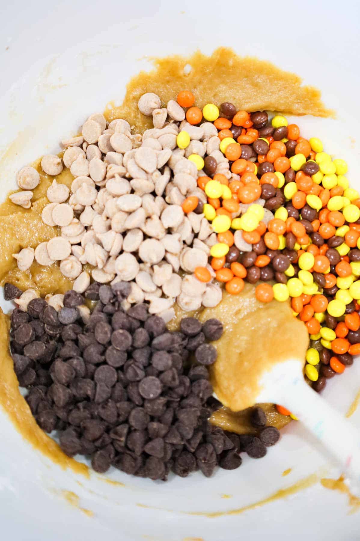 peanut butter baking chips, semi sweet chocolate chips and mini Reese's pieces candies on top of peanut butter blondie batter in a mixing bowl