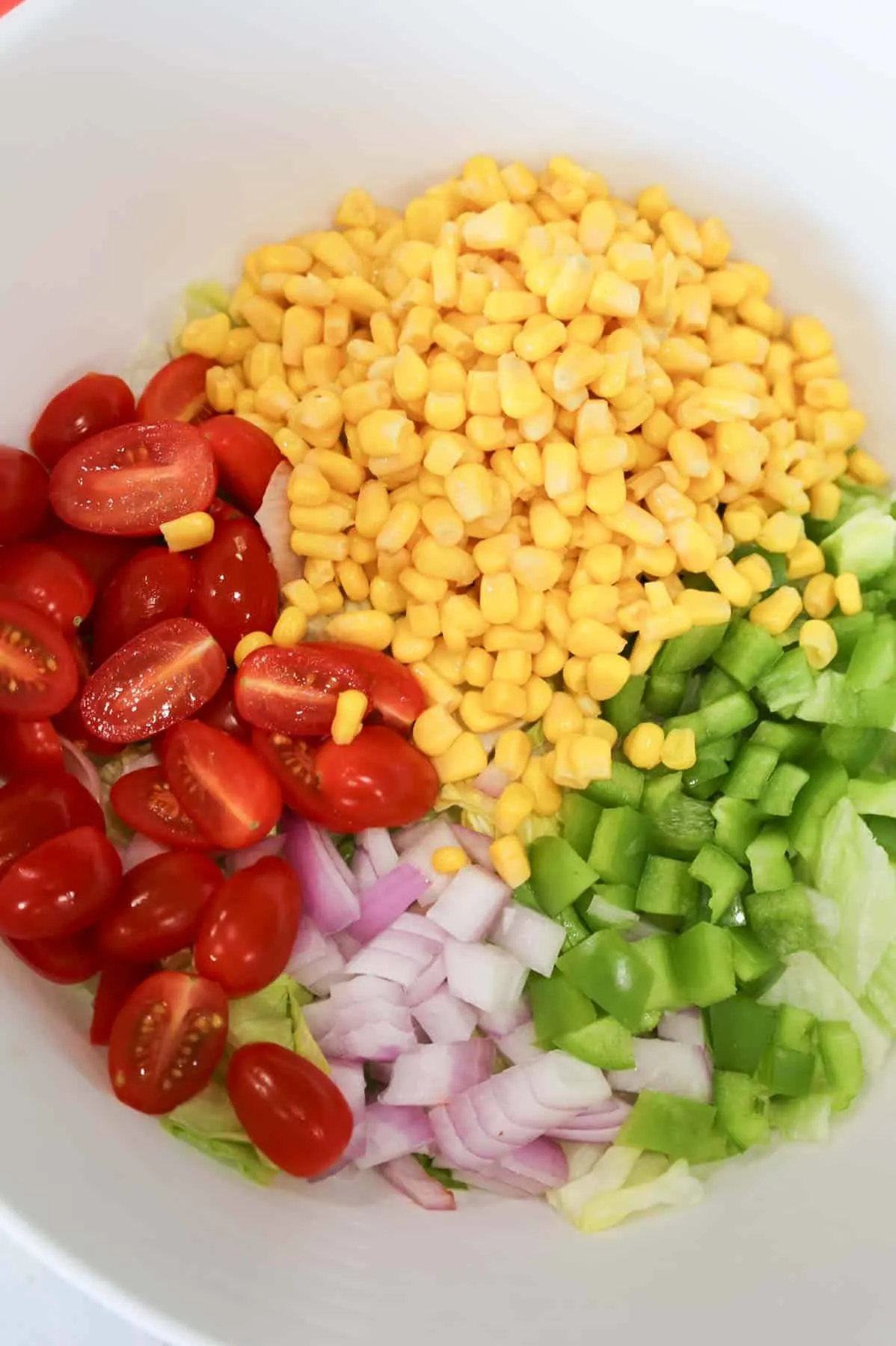 halved grape tomatoes, corn, diced green peppers and red onions on top of iceberg lettuce in a bowl