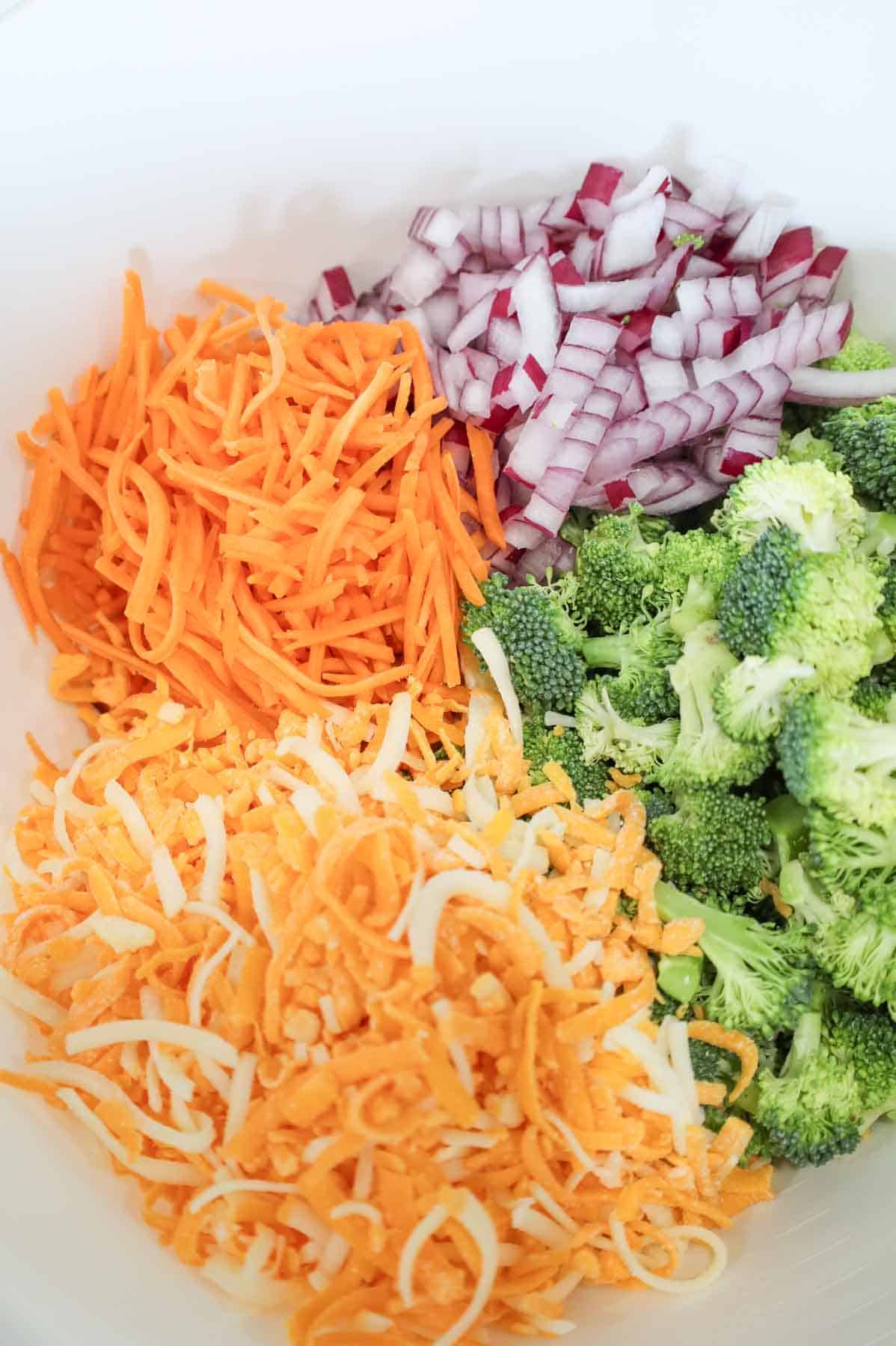 Shredded cheese, shredded carrots, diced red onions and chopped broccoli florets in a bowl