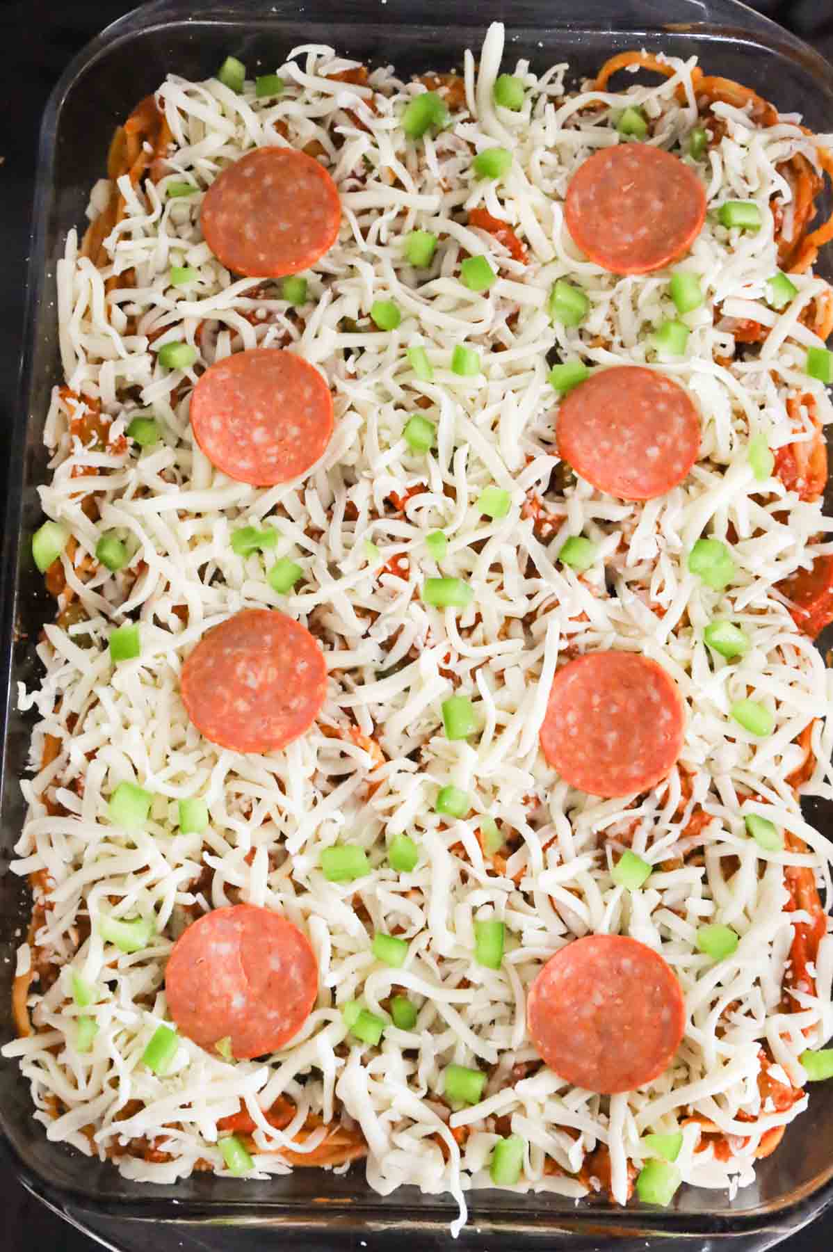 diced green peppers, pepperoni and shredded mozzarella cheese on top of spaghetti in a baking dish