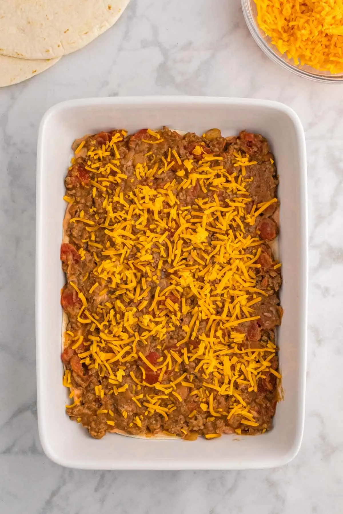 shredded cheddar cheese sprinkled over beef burrito mixture in a baking dish